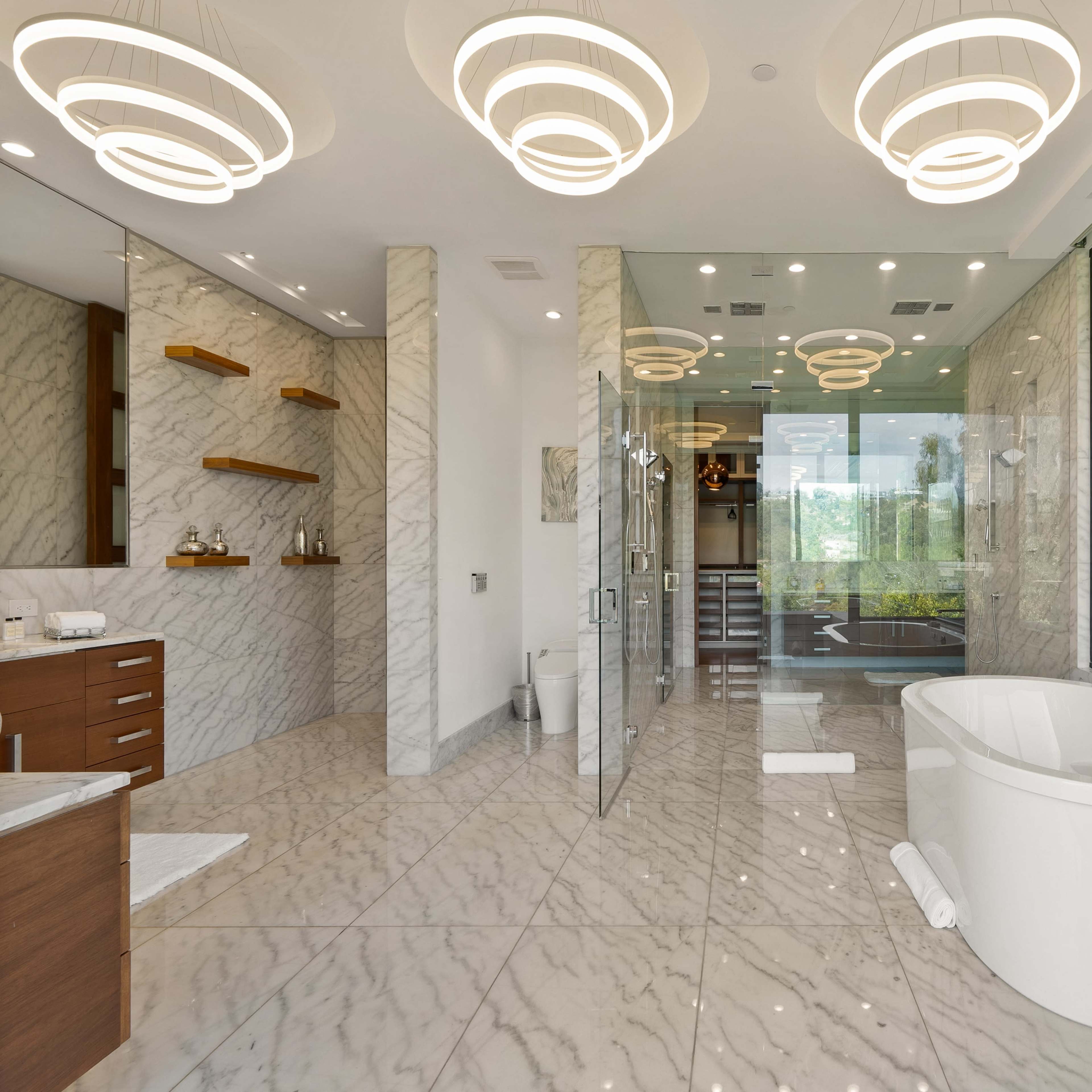 A modern bathroom featuring a freestanding bathtub, a spacious glass shower, and multiple pendant lights, all surrounded by marble walls and flooring.