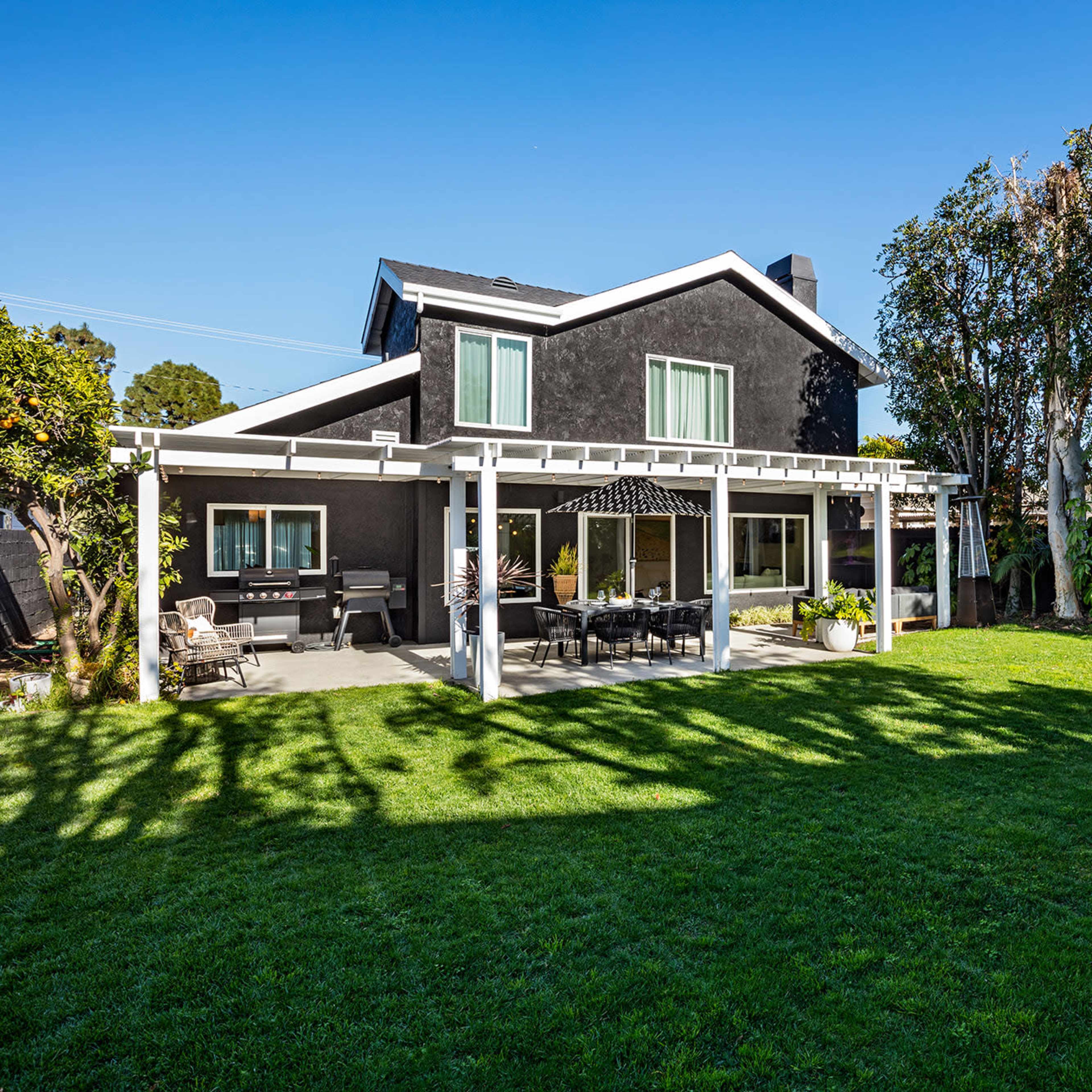 A two-story black house features a spacious backyard with a patio covered by a pergola and lush green grass.