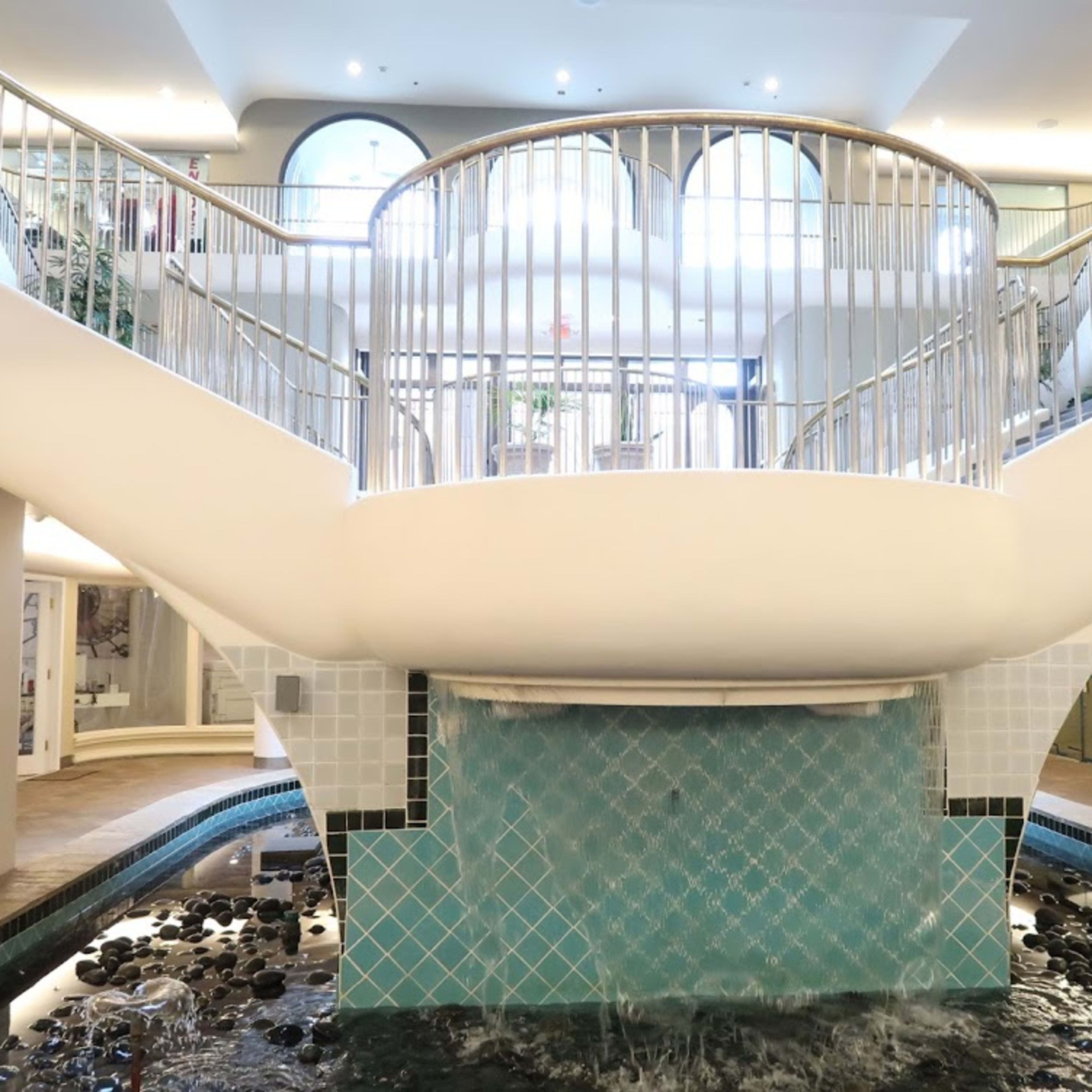 A modern interior featuring a central staircase with curved railings surrounded by a shallow water feature and tiled walls.
