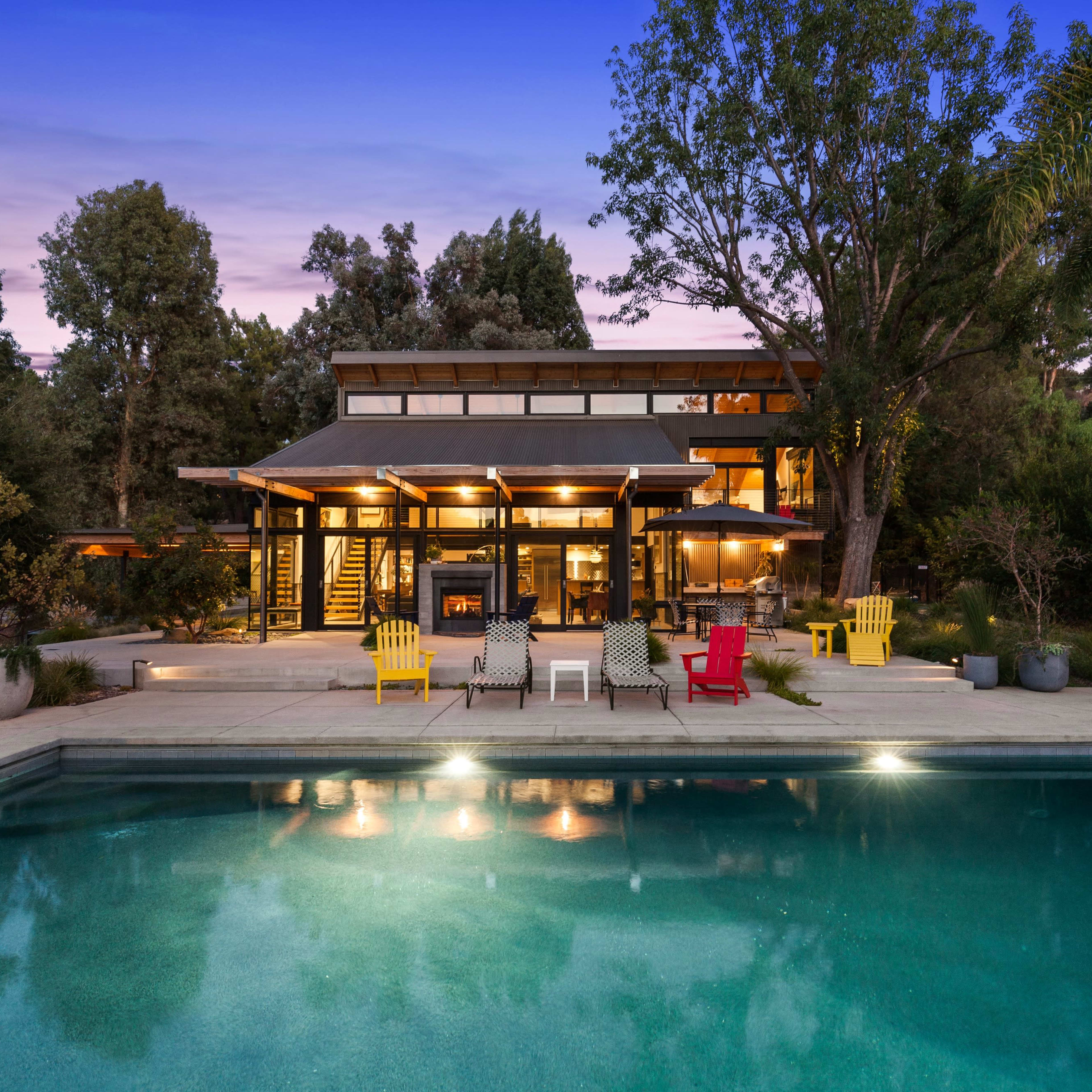 A modern house surrounded by trees features a swimming pool in the foreground and outdoor seating areas with colorful chairs.