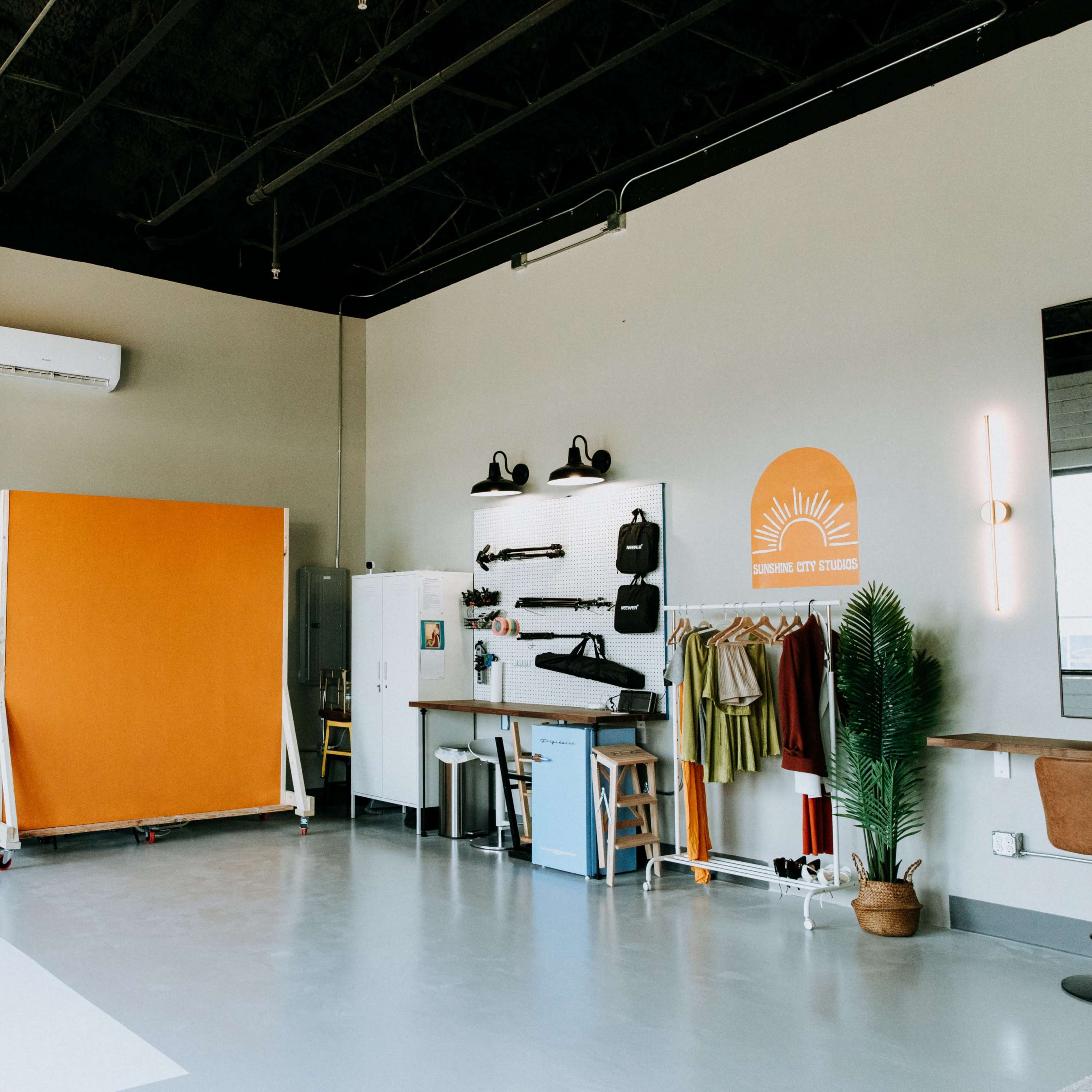 A photography studio with a white backdrop, an orange backdrop, various photographic equipment, a wardrobe rack, and a small seating area.