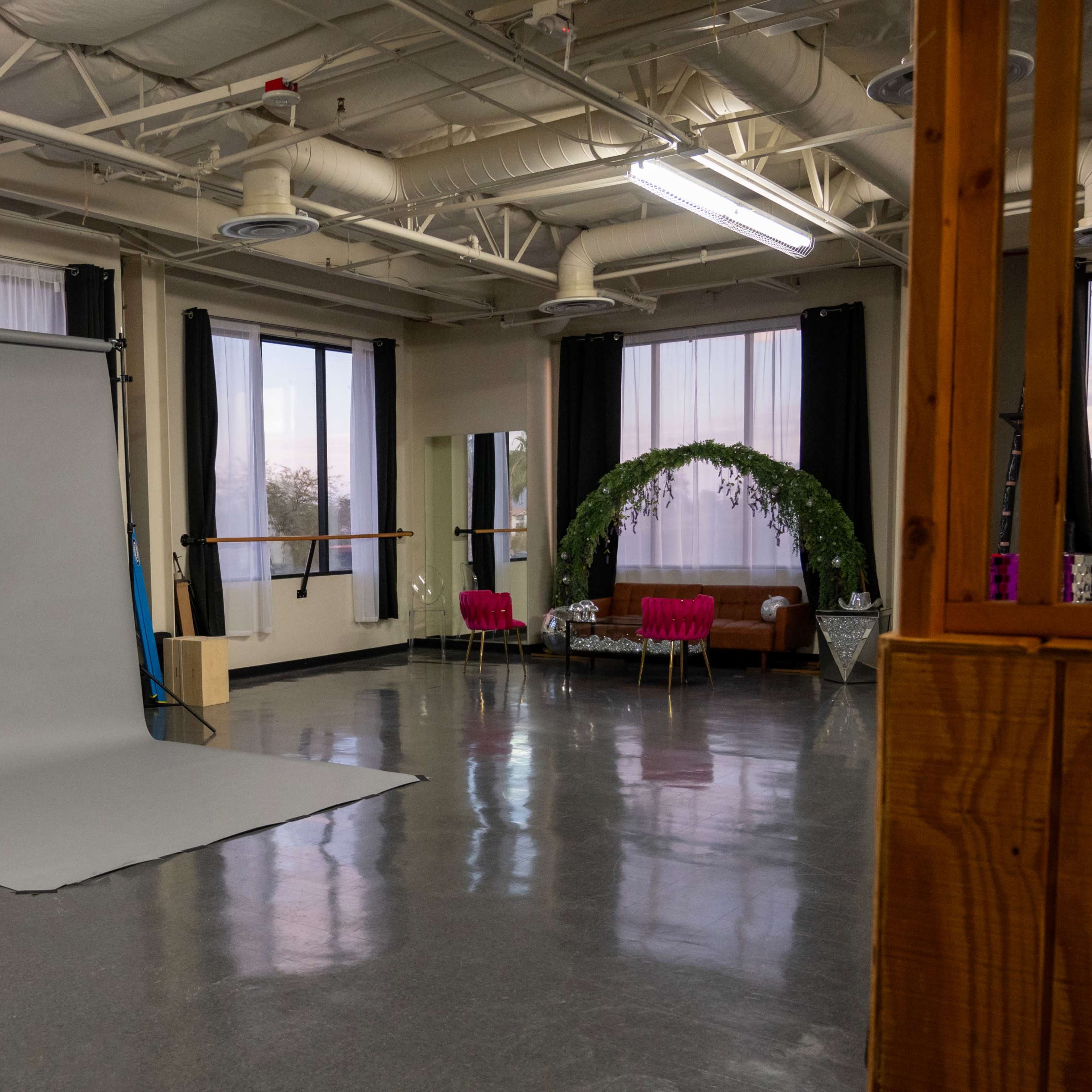 A spacious studio with a gray backdrop, large windows, and a seating area featuring pink chairs and greenery.