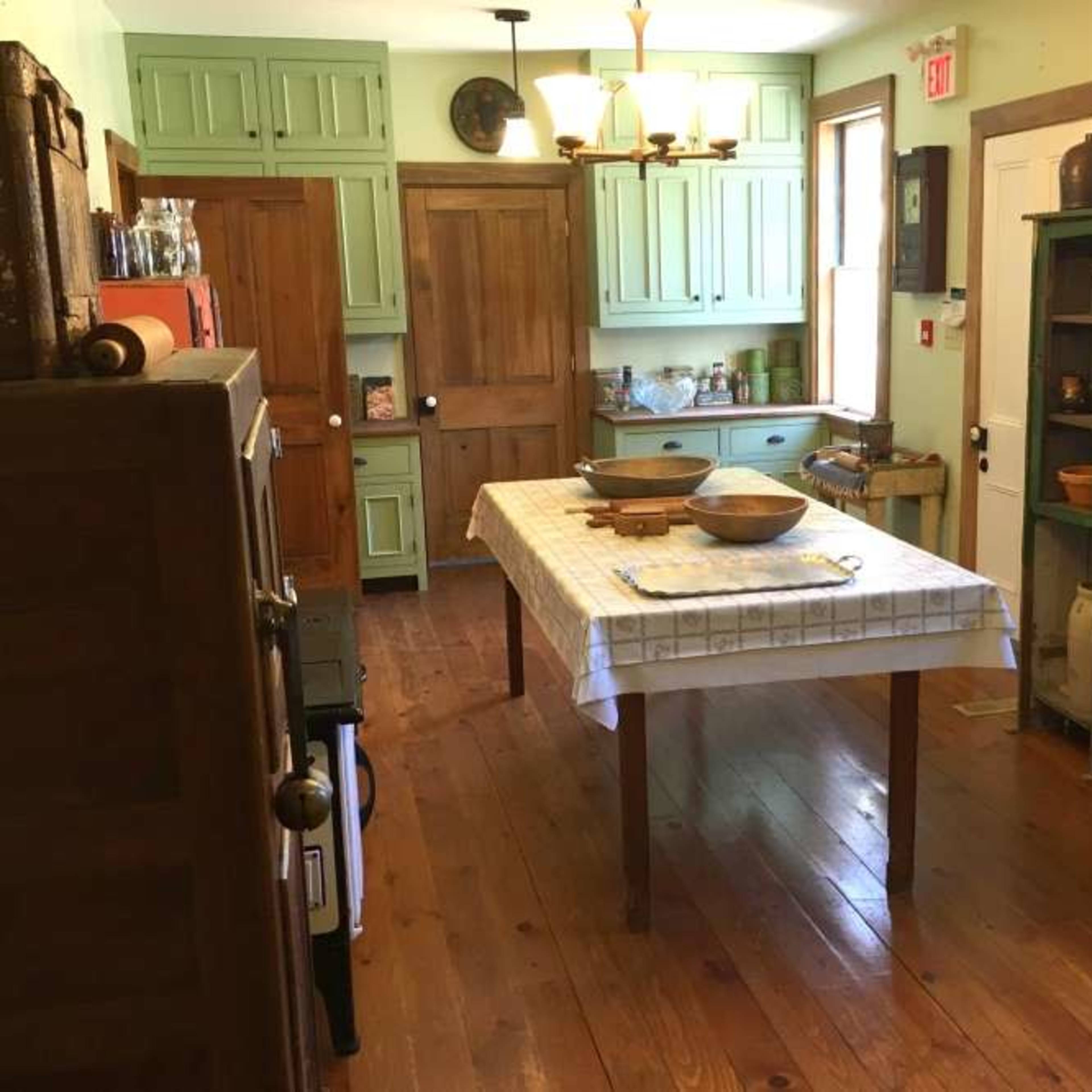 A quaint kitchen with wooden floors, a central table, and green cabinetry along the walls.