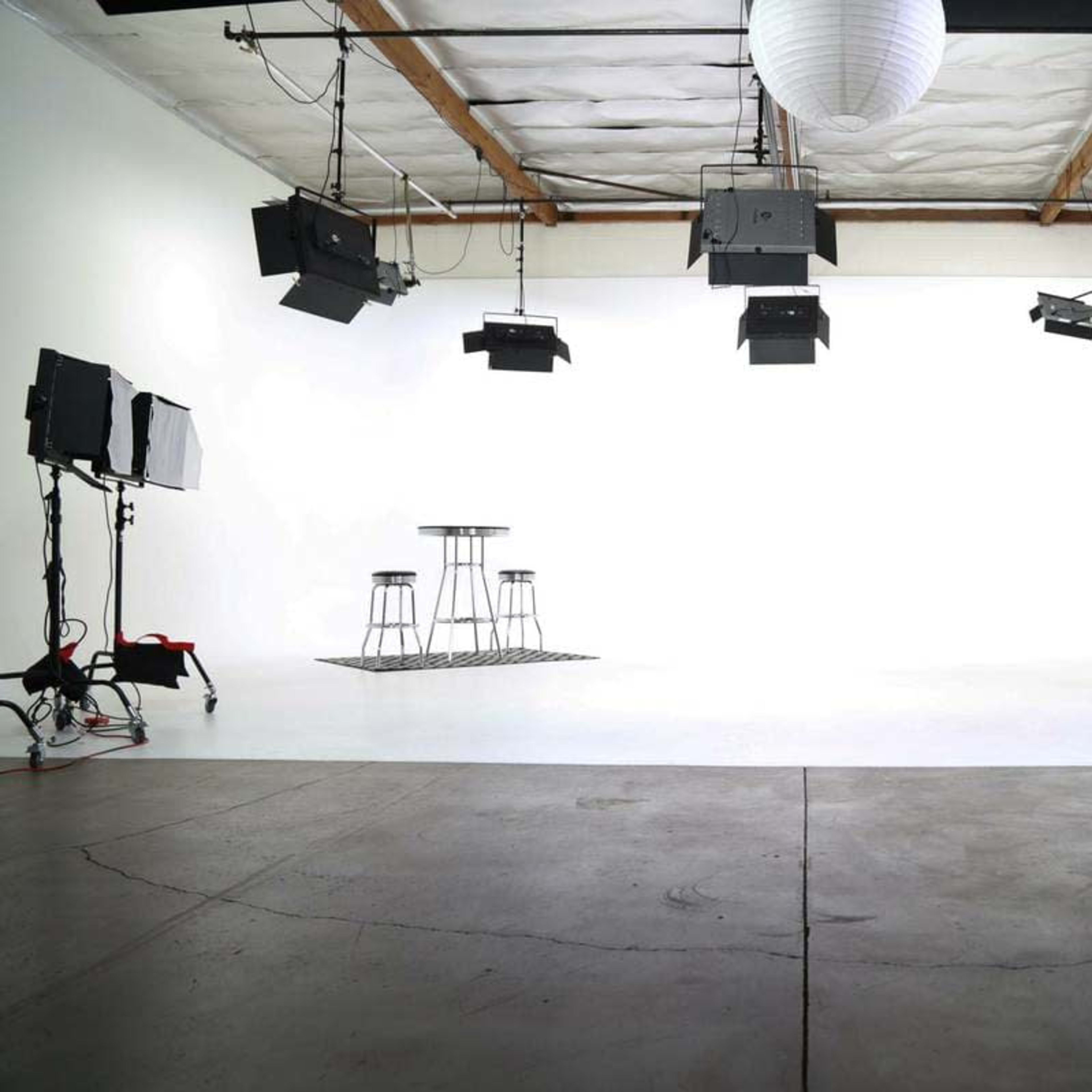 A minimally furnished studio space with two bar stools, a table, and various lighting equipment suspended from the ceiling.