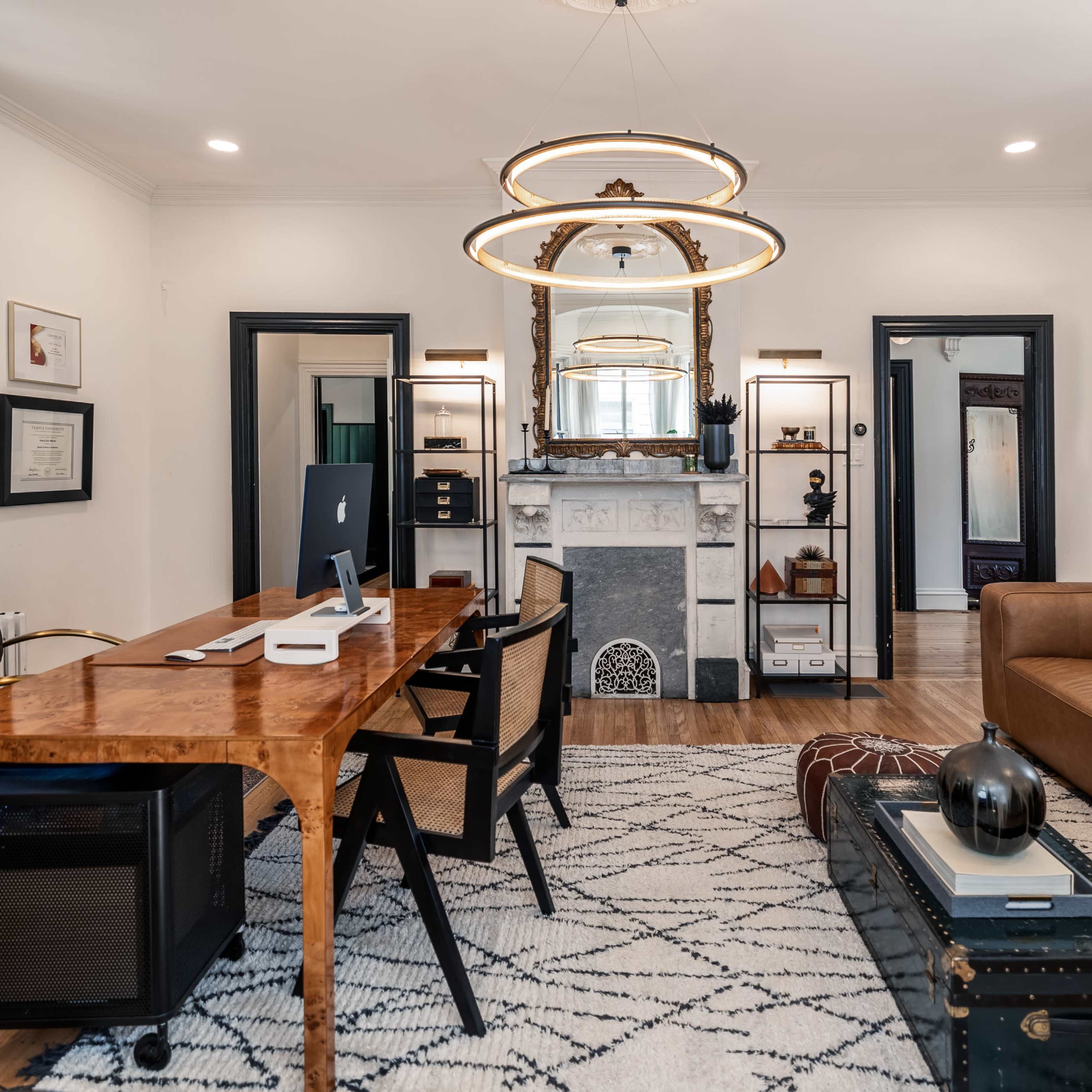 A modern office features a large wooden desk, a brown sectional couch, a marble fireplace with a mirror above, and a patterned rug on the wooden floor.