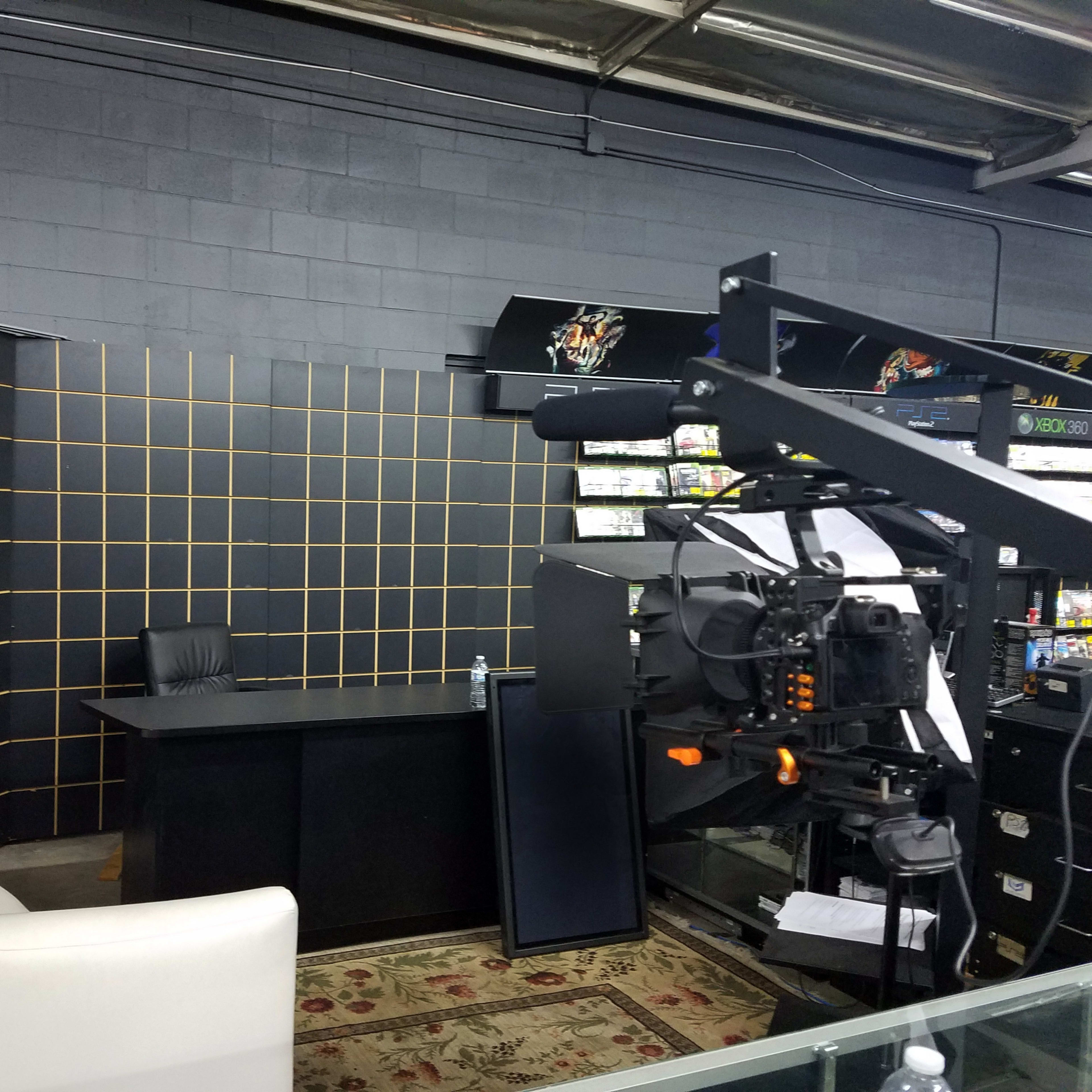 A video production setup featuring a white couch, a desk, and a camera on a tripod against a backdrop of black and orange grid patterns.
