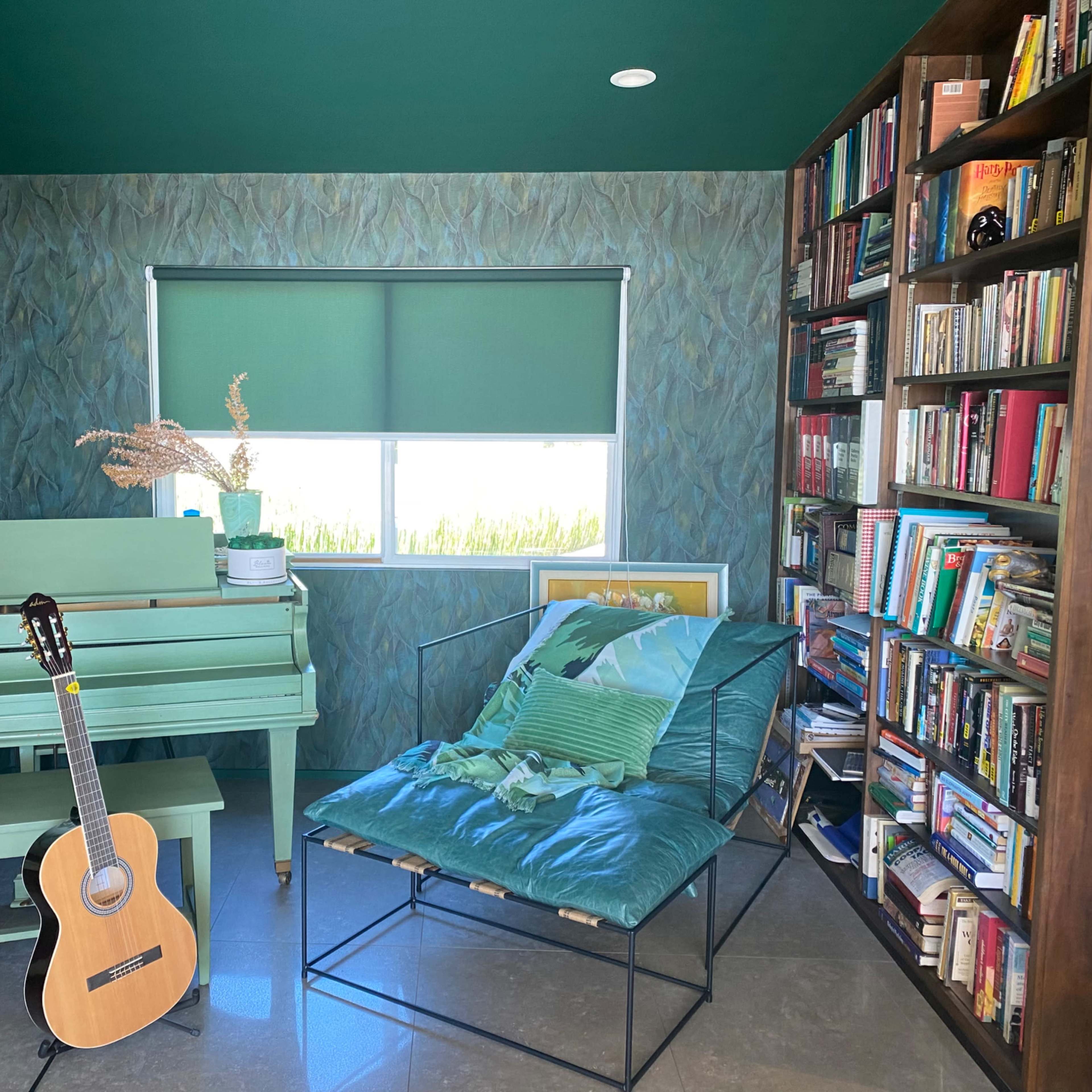 A green-themed room features a piano, a guitar, a lounge chair, and a bookshelf filled with books.
