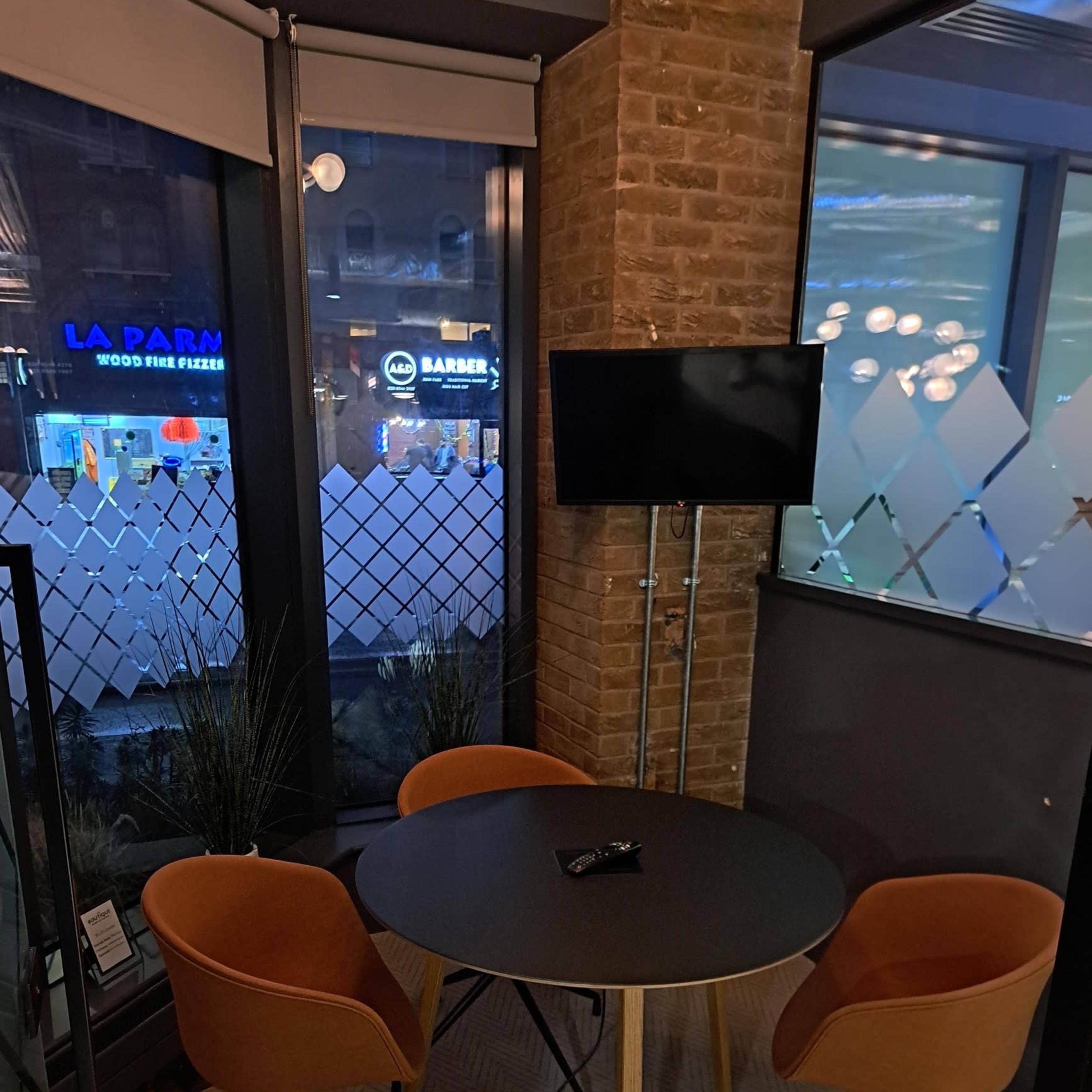 A small circular table with two orange chairs and a wall-mounted TV in a corner office space with large windows.