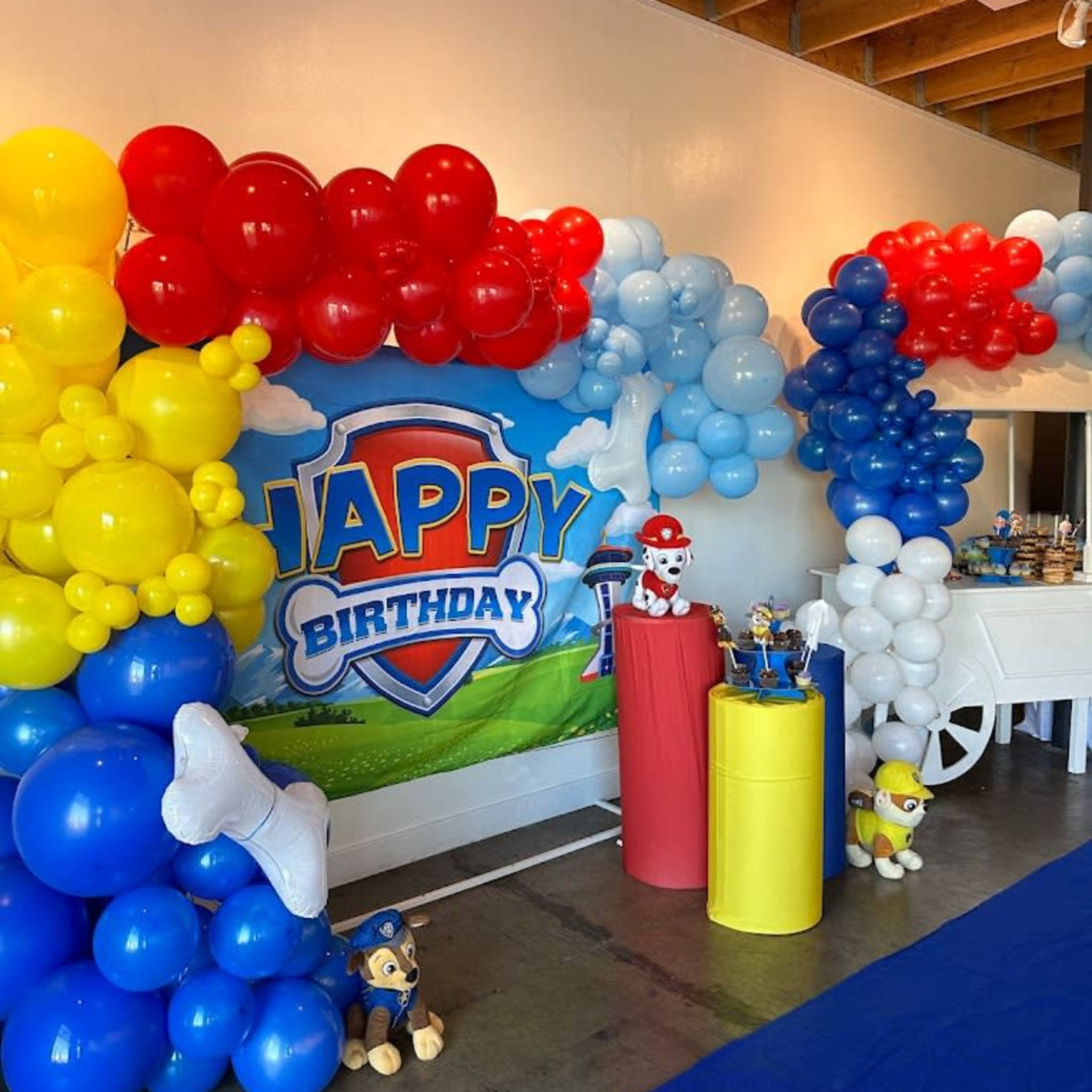 A colorful birthday party setup featuring balloon decorations in red, blue, yellow, and white, along with a backdrop that says "Happy Birthday" and some cartoon characters displayed on tables.