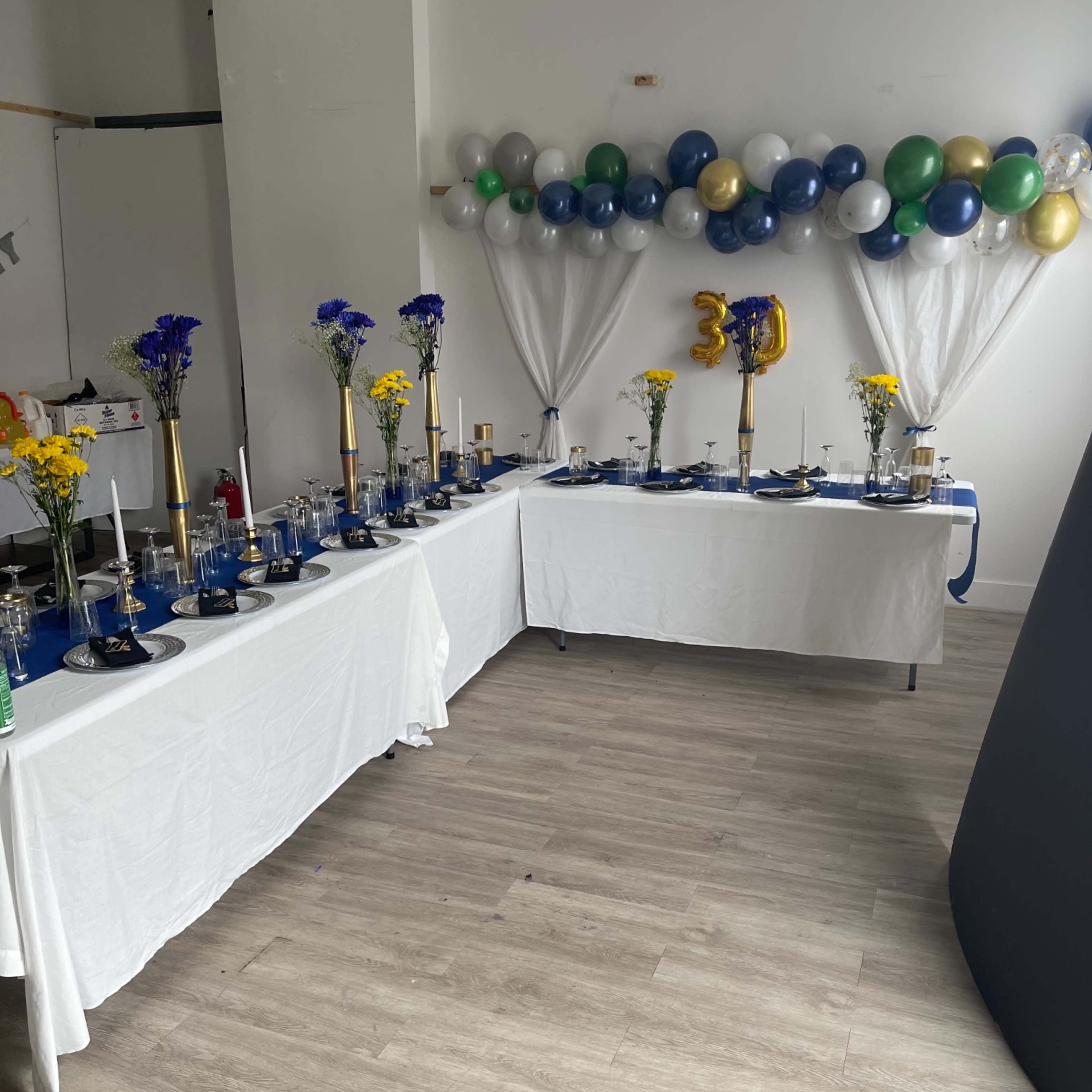 A decorated party setup with two tables covered in white cloths, featuring blue and gold centerpieces, and a balloon arch in the background celebrating a 30th birthday.