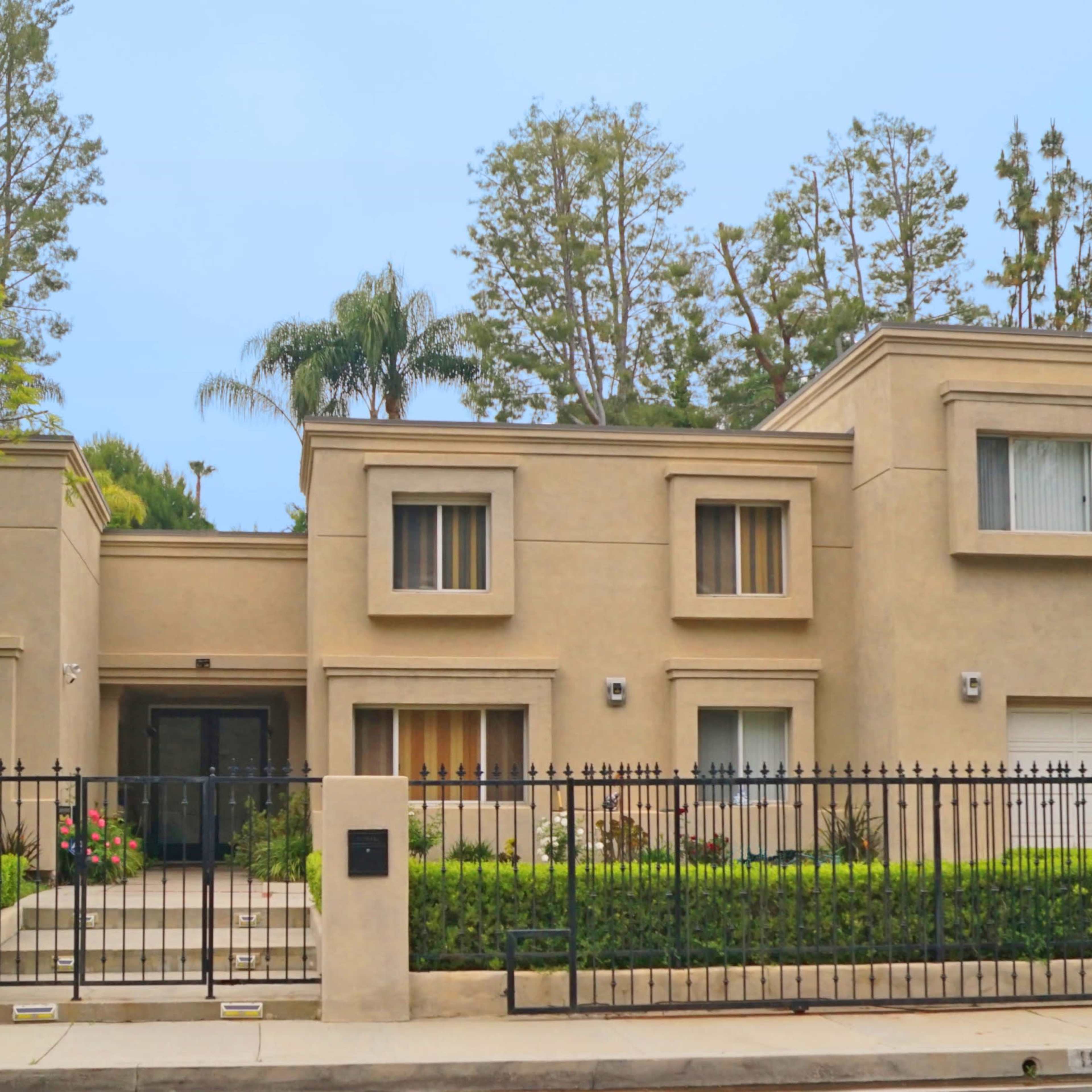 A large, modern, beige house surrounded by greenery and enclosed by a black wrought-iron fence.