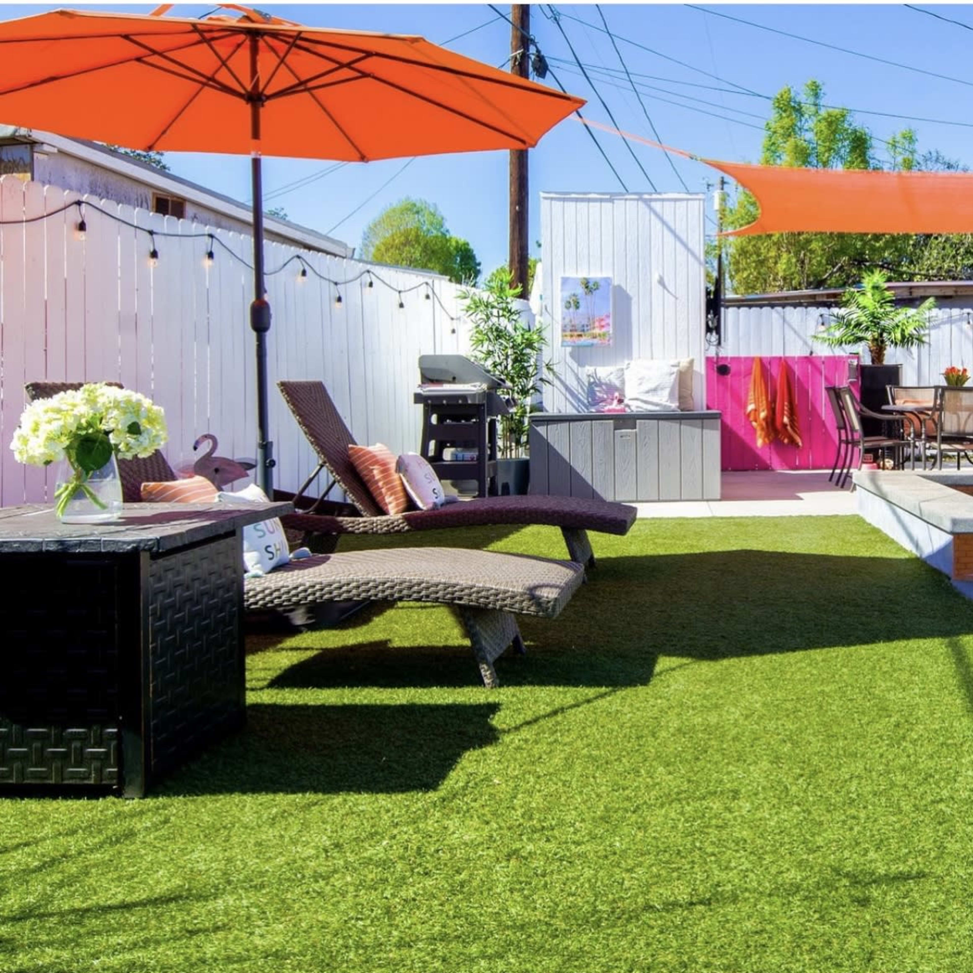 A landscaped backyard with two lounge chairs, a table, a grill, and colorful umbrellas, surrounded by white fencing and greenery.