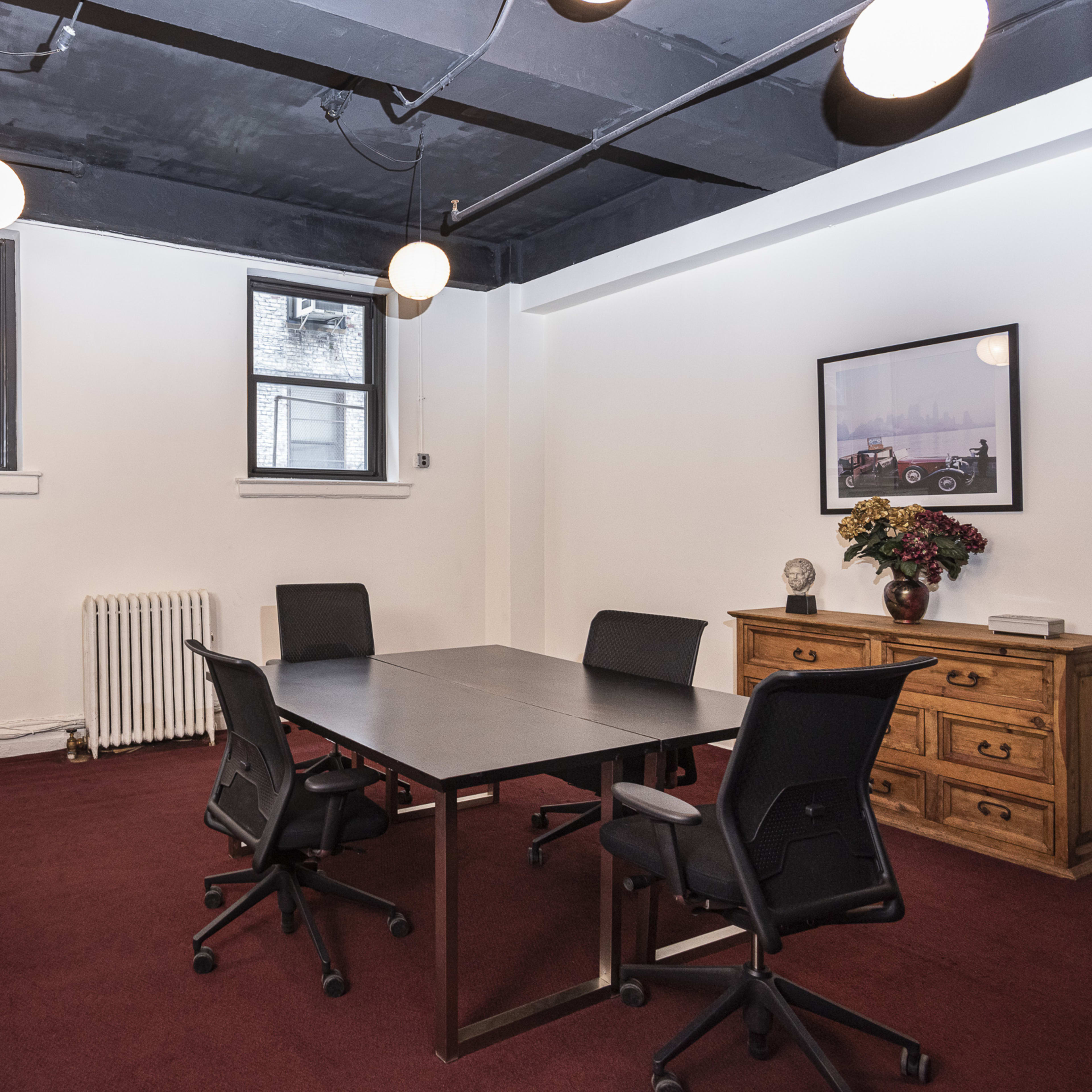 A conference room features a rectangular table surrounded by four chairs, with windows, a cabinet, and a decorative photo on the wall.