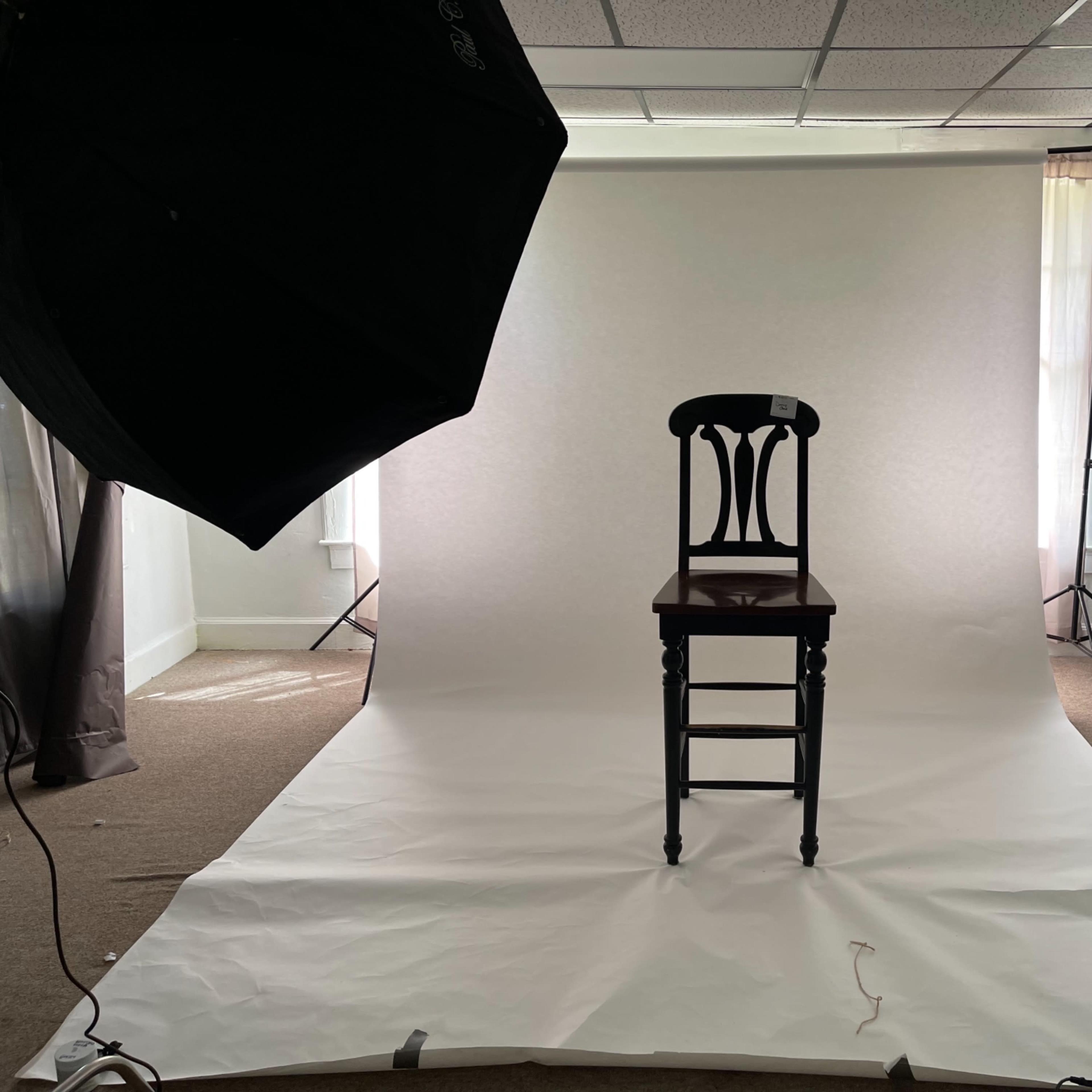 A black chair is centered in a brightly lit studio surrounded by white backdrop paper and photography equipment.