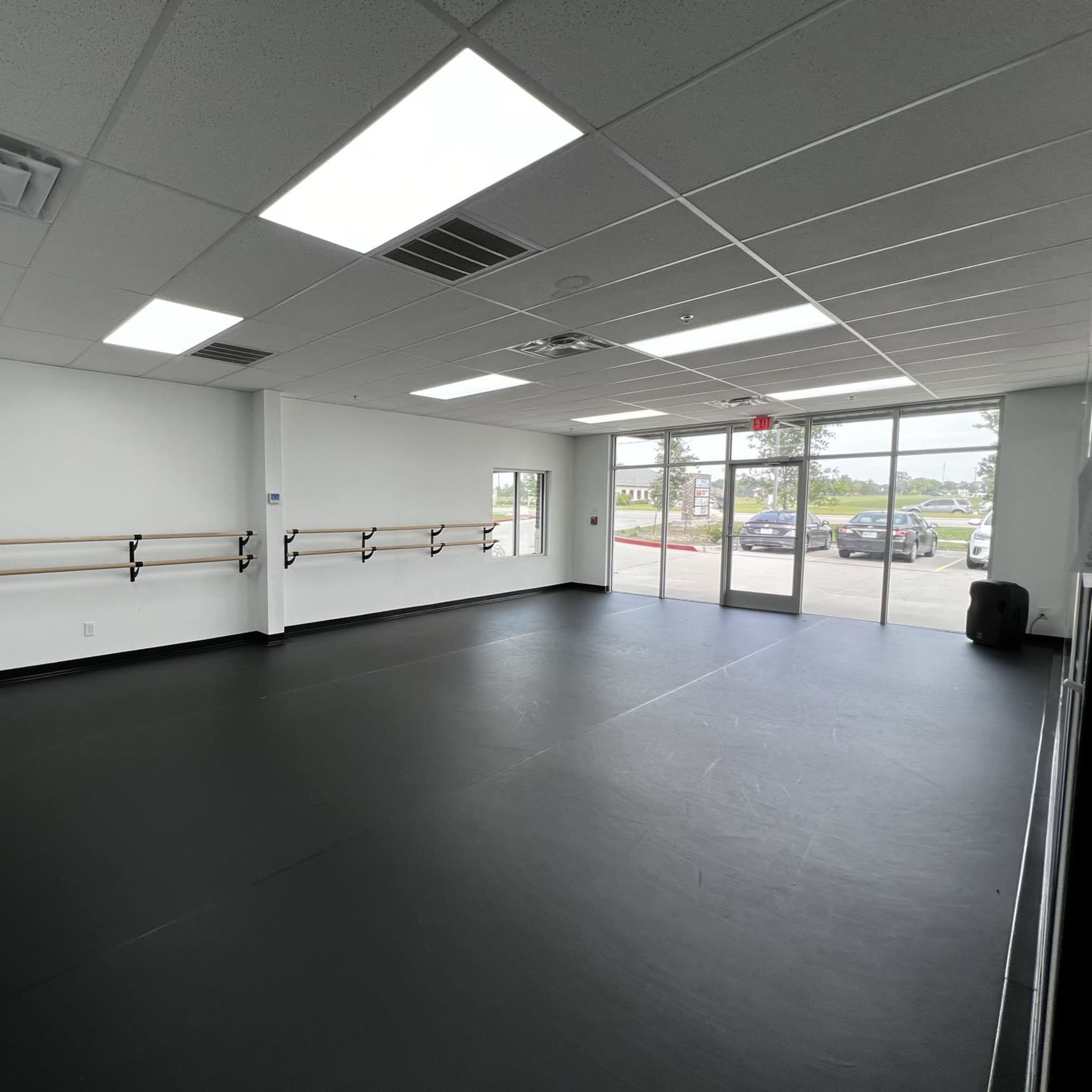 A spacious dance studio with a black floor, mirrored walls, and ballet barres along one side, along with large windows letting in natural light.