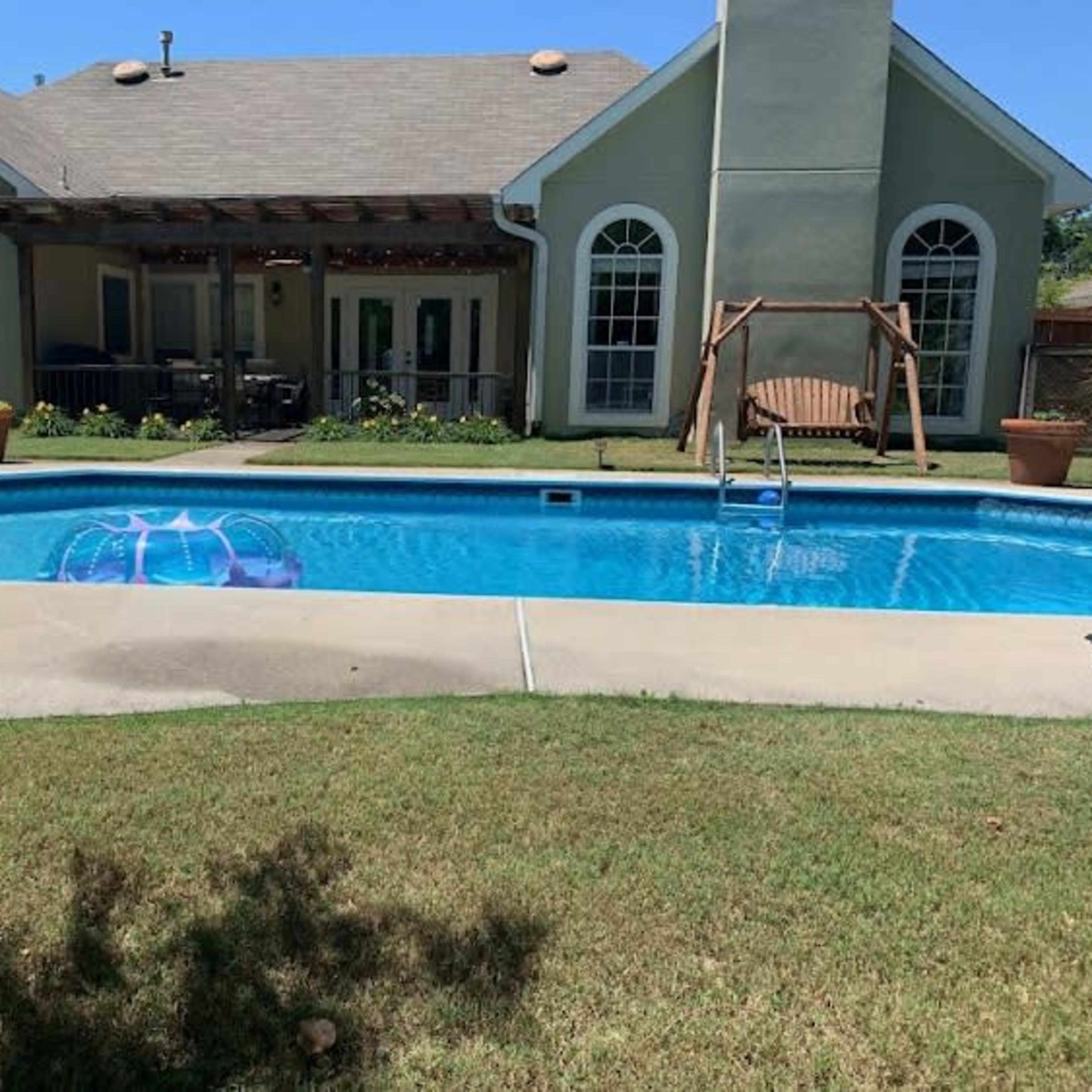 A backyard with a swimming pool, a house in the background, and a swing set nearby.
