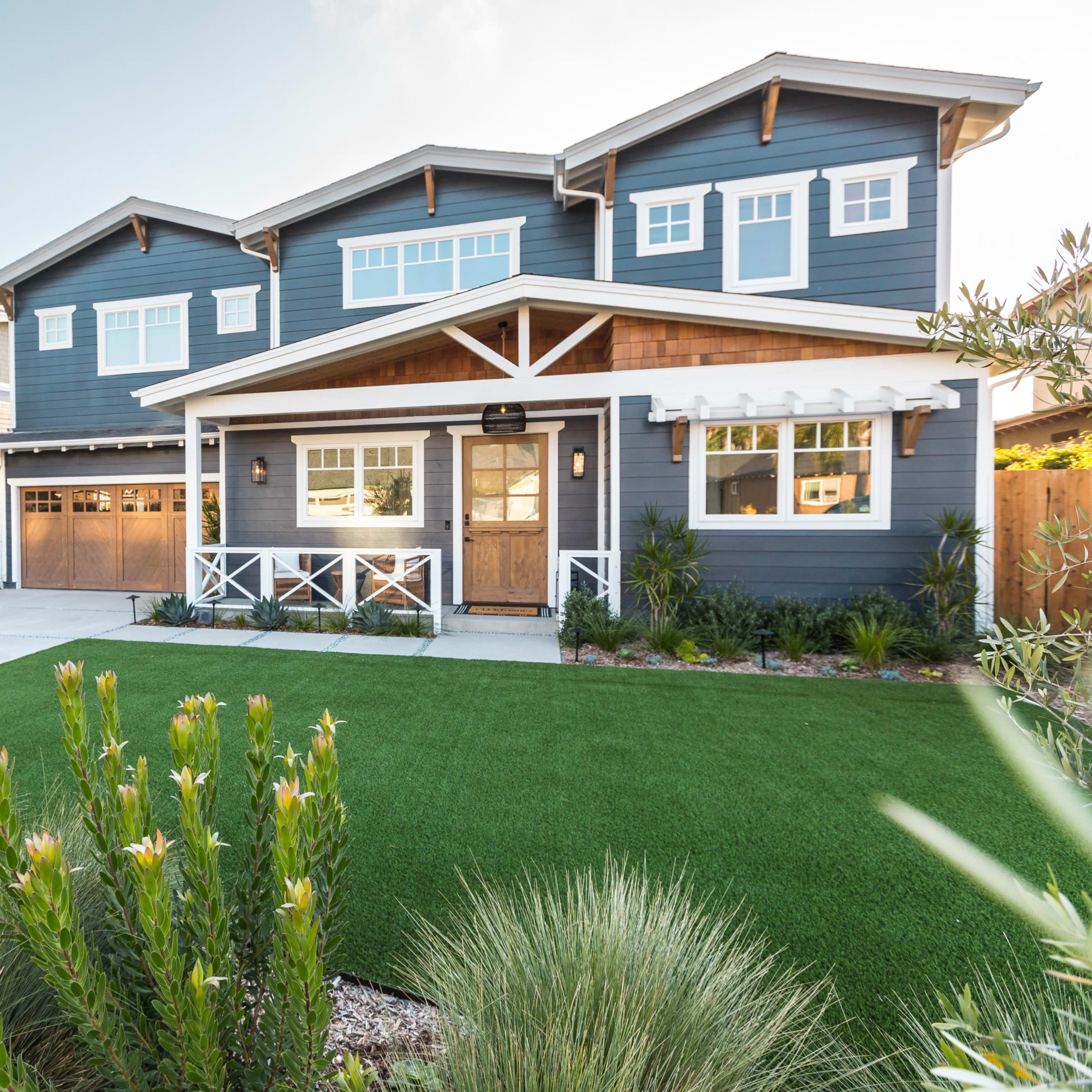 A two-story blue house with a wooden porch and garage doors is surrounded by landscaped greenery and artificial turf.