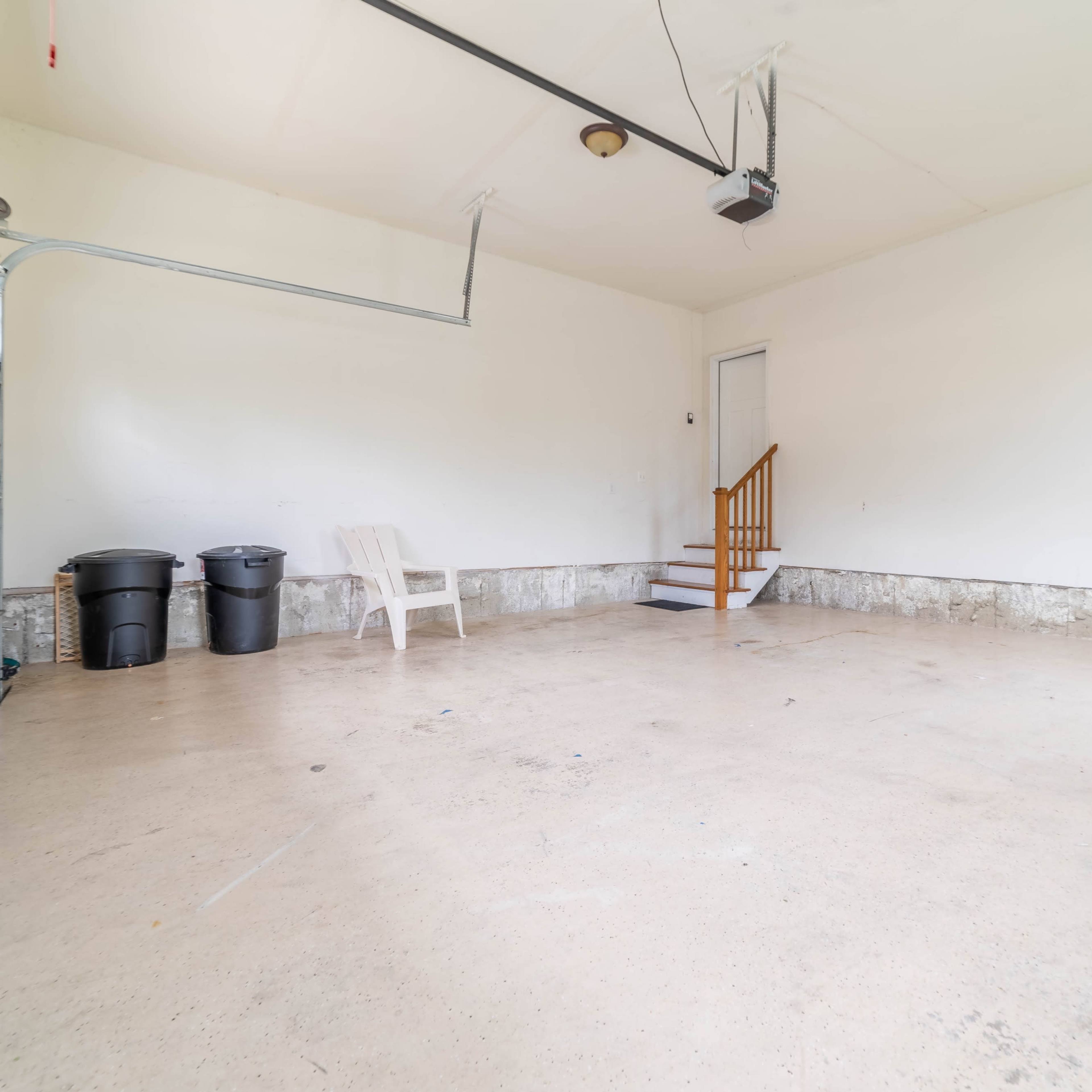 An empty garage with a concrete floor, two trash bins, a plastic chair, and a set of stairs leading to a door.
