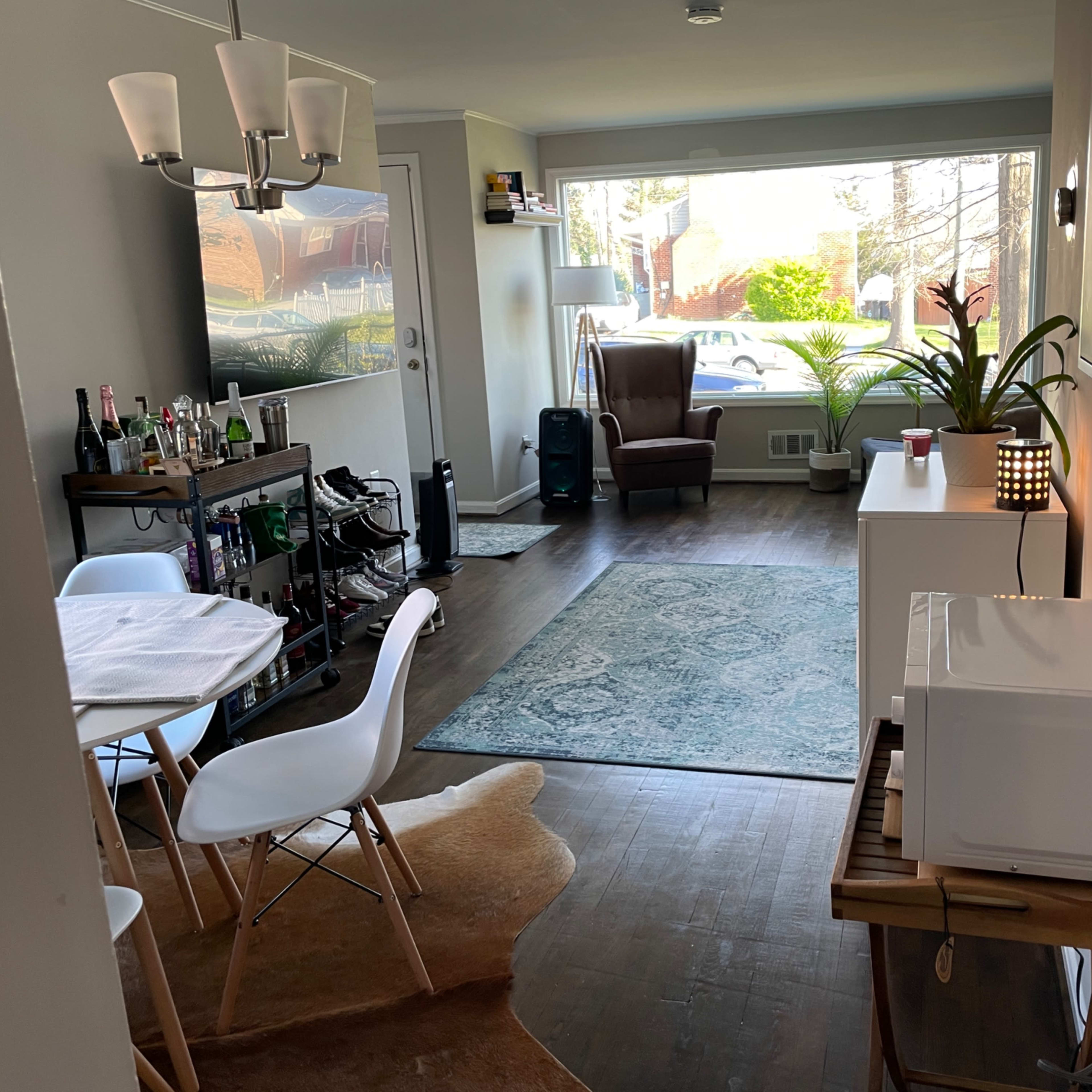 A hallway leads to a living area with a dining table, a large window, and a comfortable chair, featuring a plant and a microwave.
