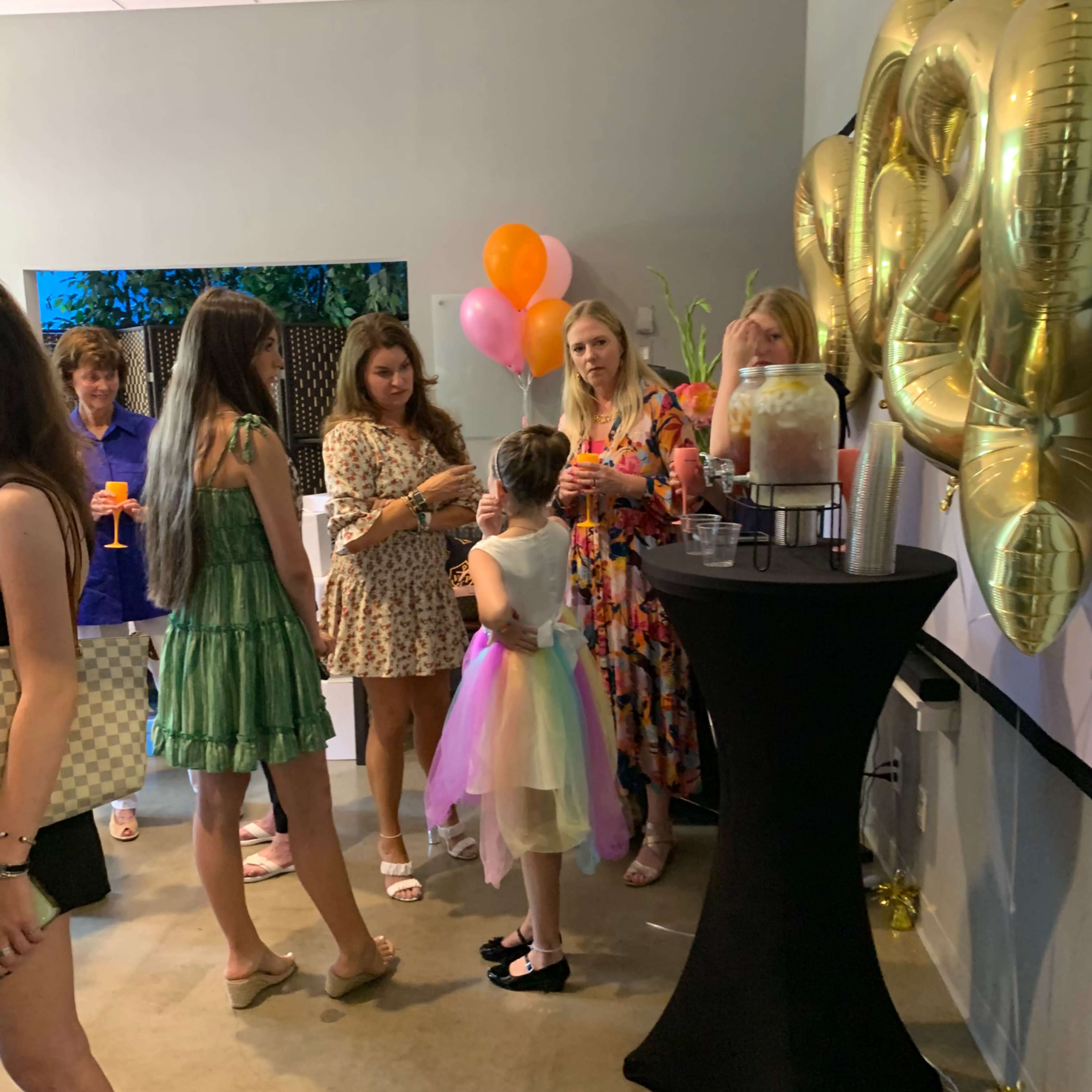 A group of women and a child in a colorful outfit gather around a table with drinks and decorations at a celebration.