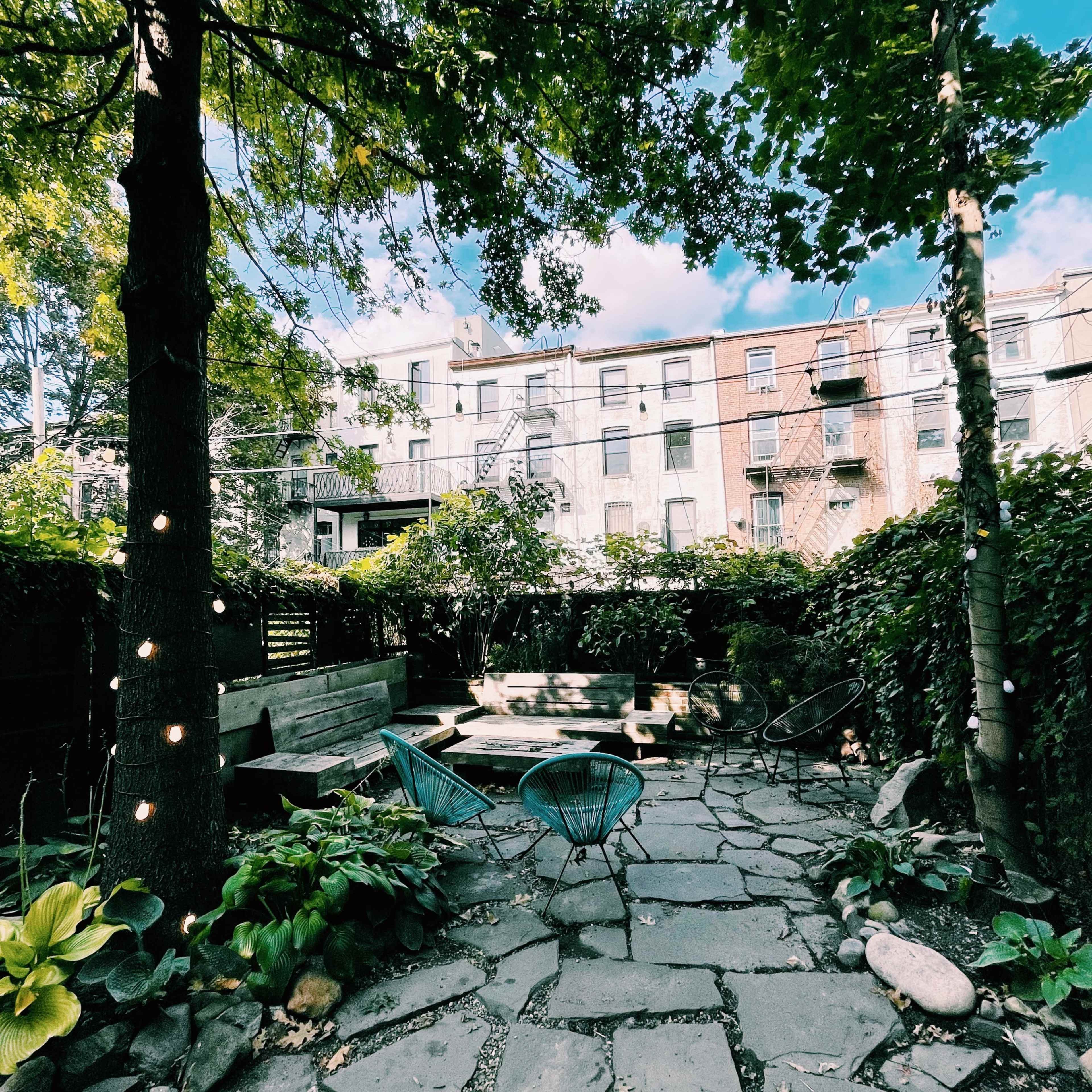 A stone-paved backyard garden with green foliage, trees, and a building with balconies in the background.