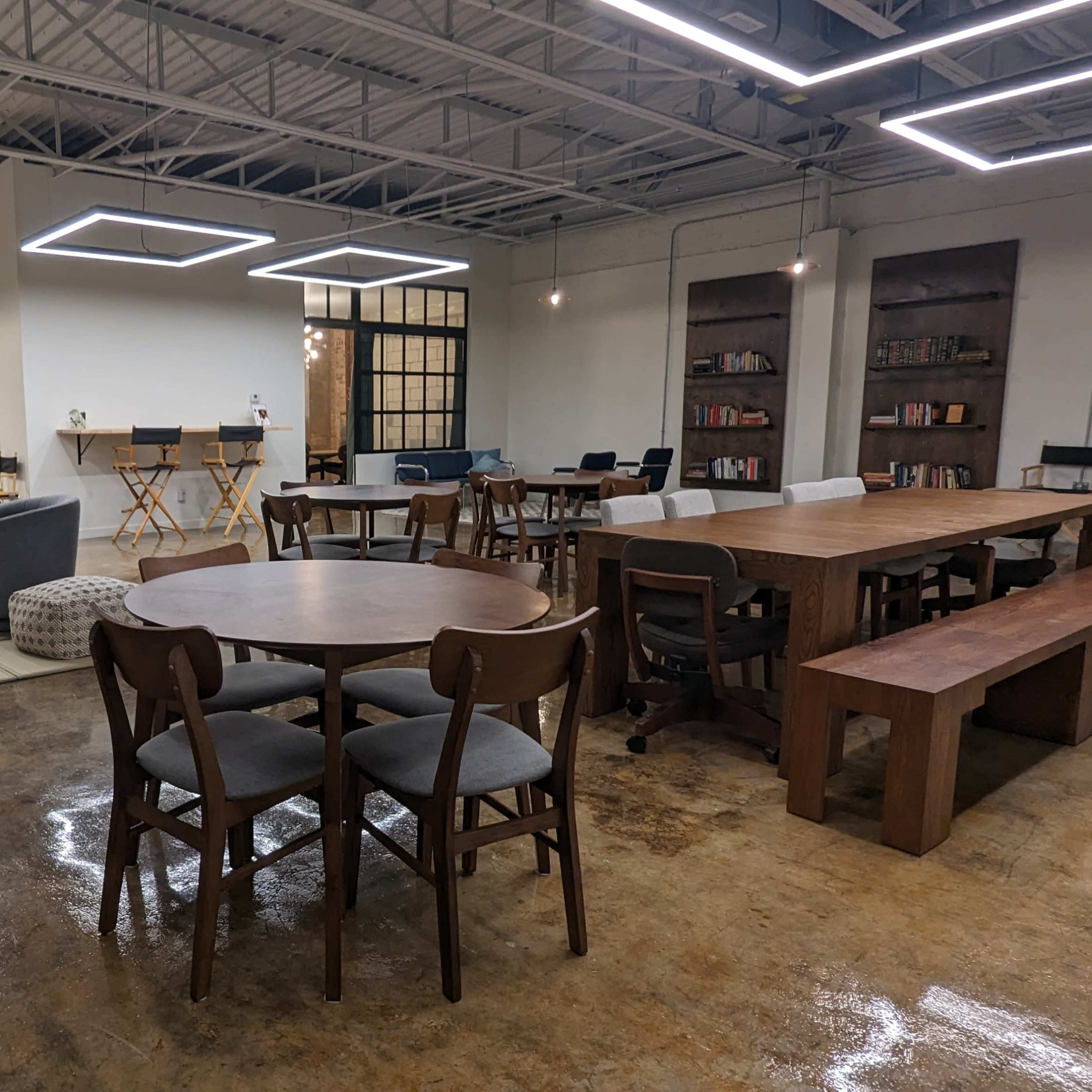 A modern, well-lit co-working space with various seating arrangements, including tables, chairs, and couches, along with shelves of books.
