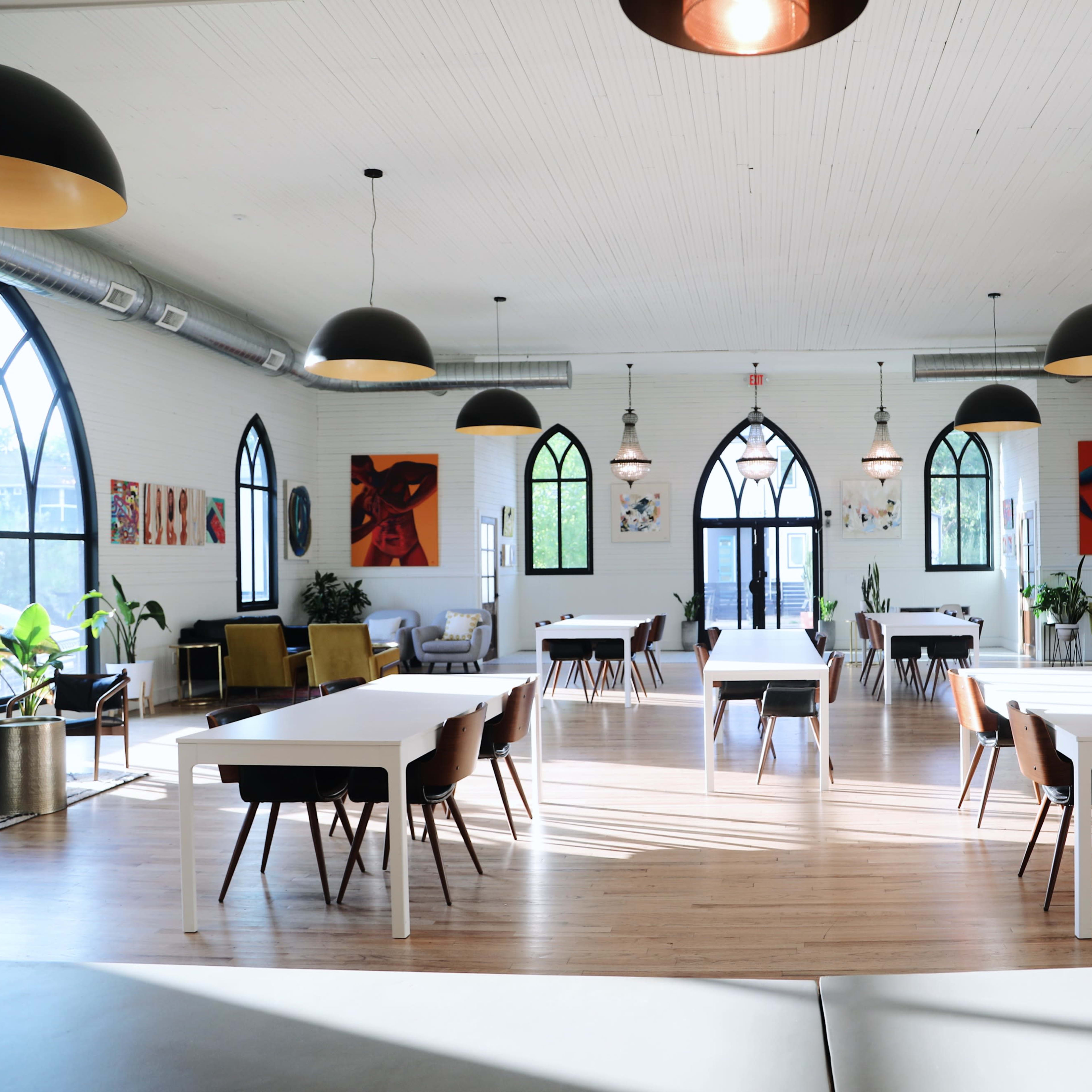 A spacious, well-lit interior of a room with high ceilings, featuring tables and chairs, large arched windows, and various artworks on the walls.