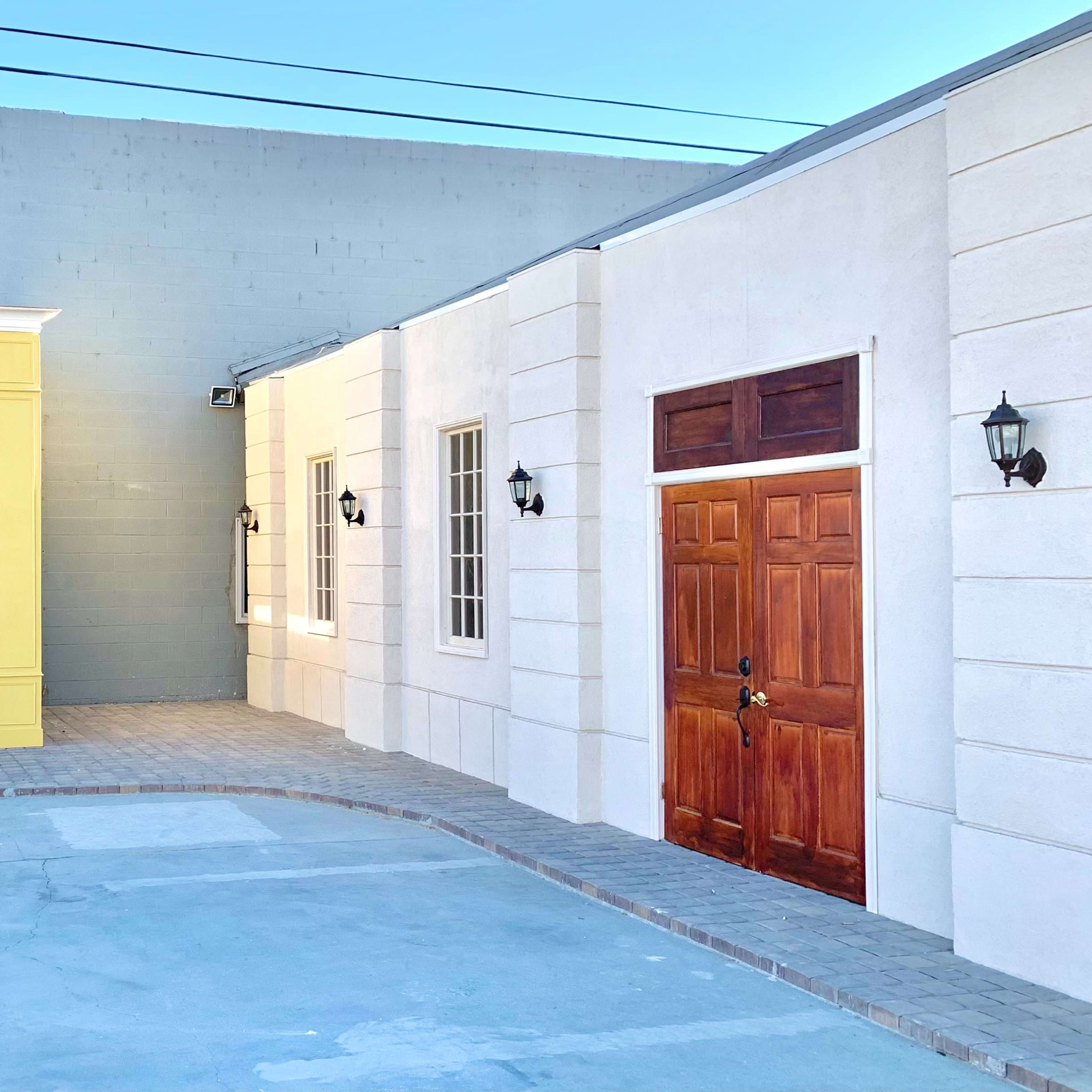 A corner of a building complex with a yellow storefront on the left and a white building with a wooden door on the right.