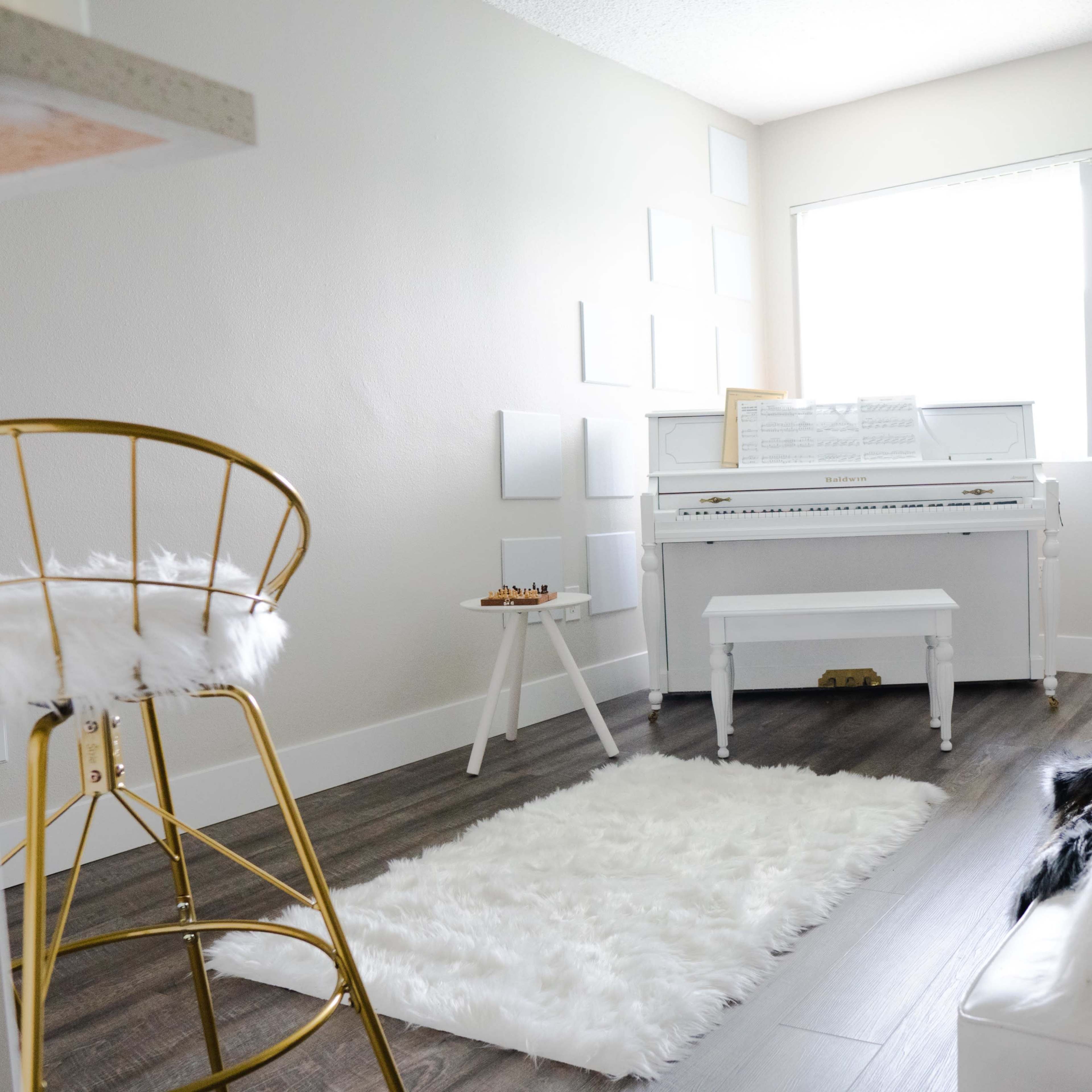 A bright room features a white piano, a plush rug, a gold bar stool, and a sofa with decorative pillows.