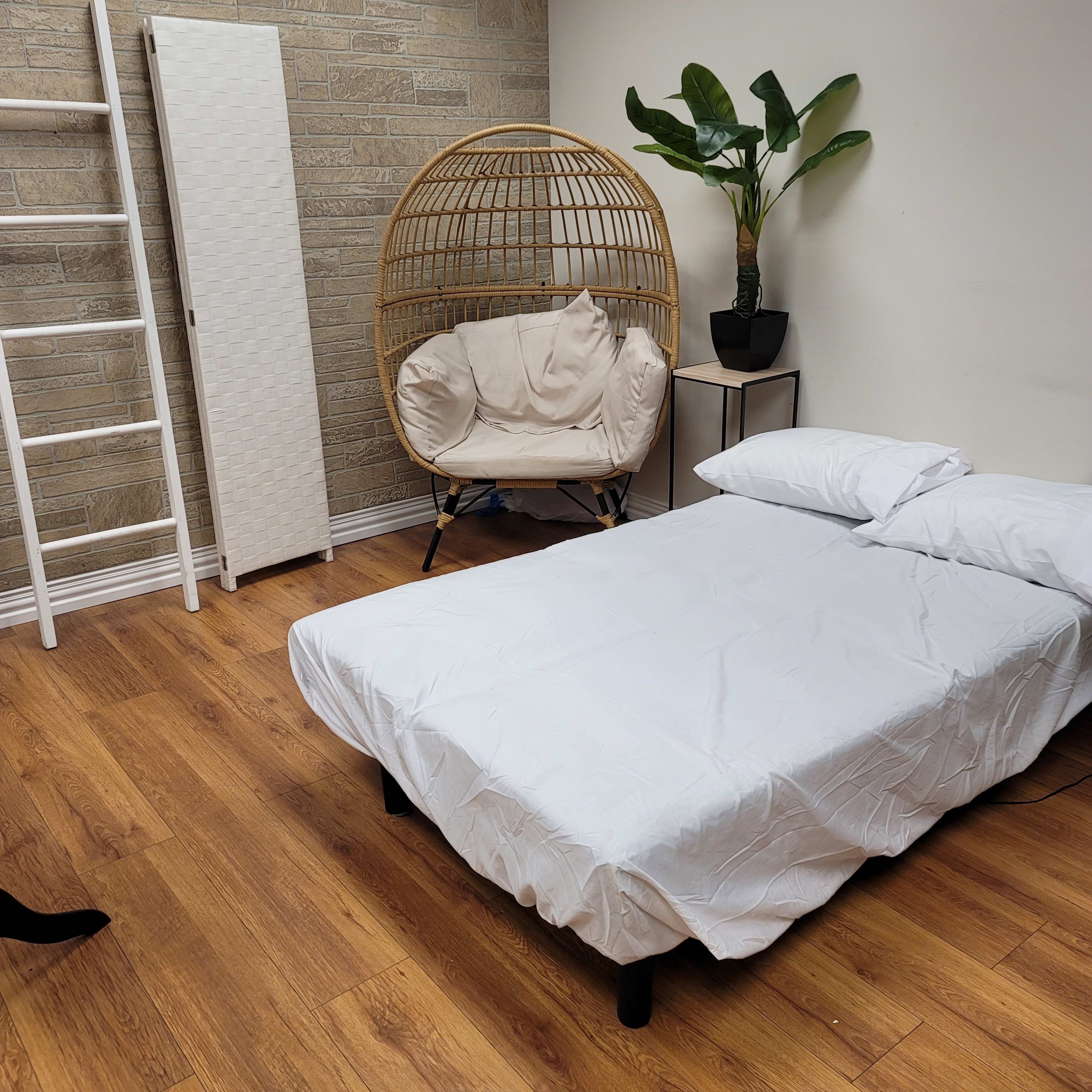 A bedroom with a bed covered in a white sheet, a round wicker chair, a potted plant, and a white folding screen against a textured wall.