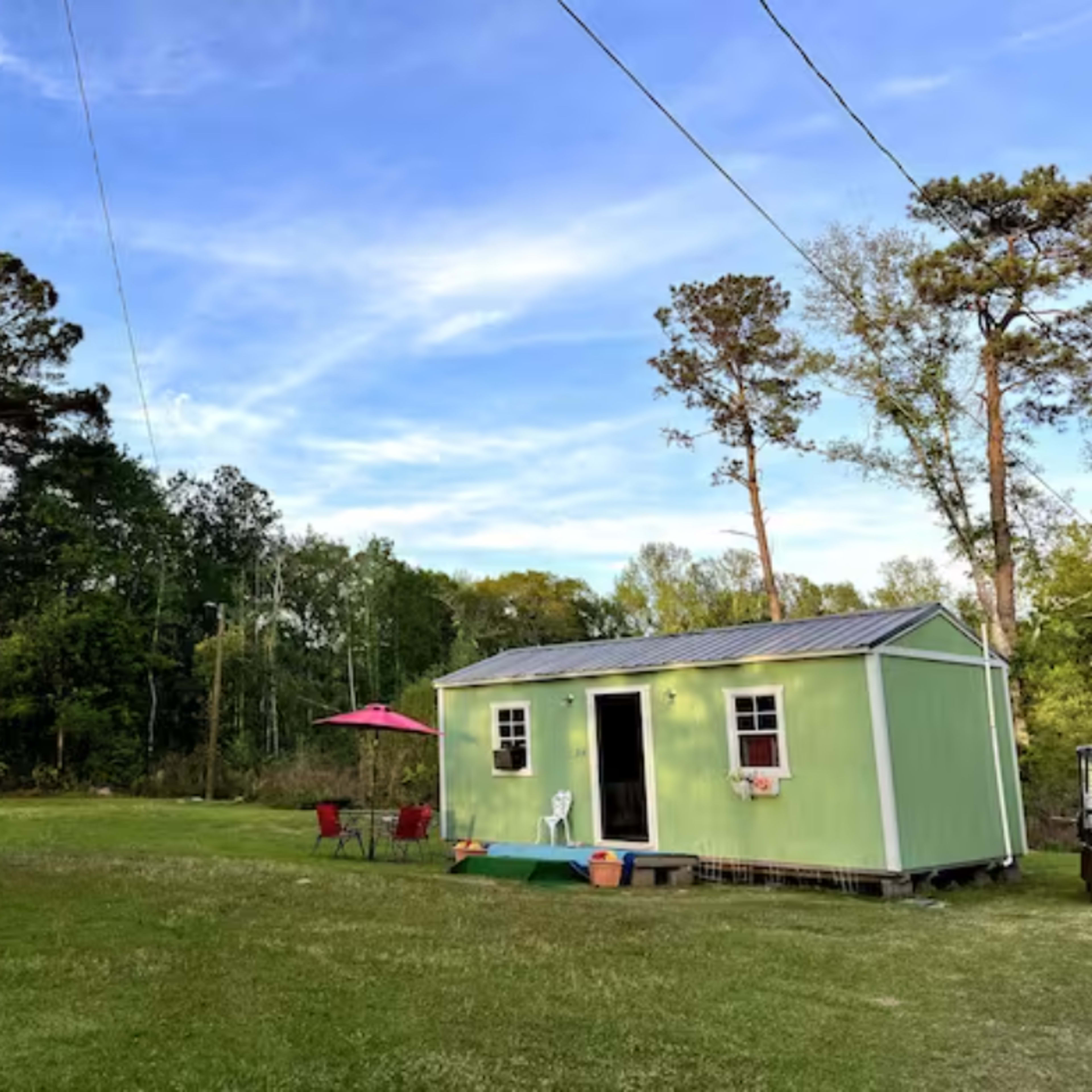 A small green house with a metal roof is situated in a grassy area, surrounded by trees and power lines above.