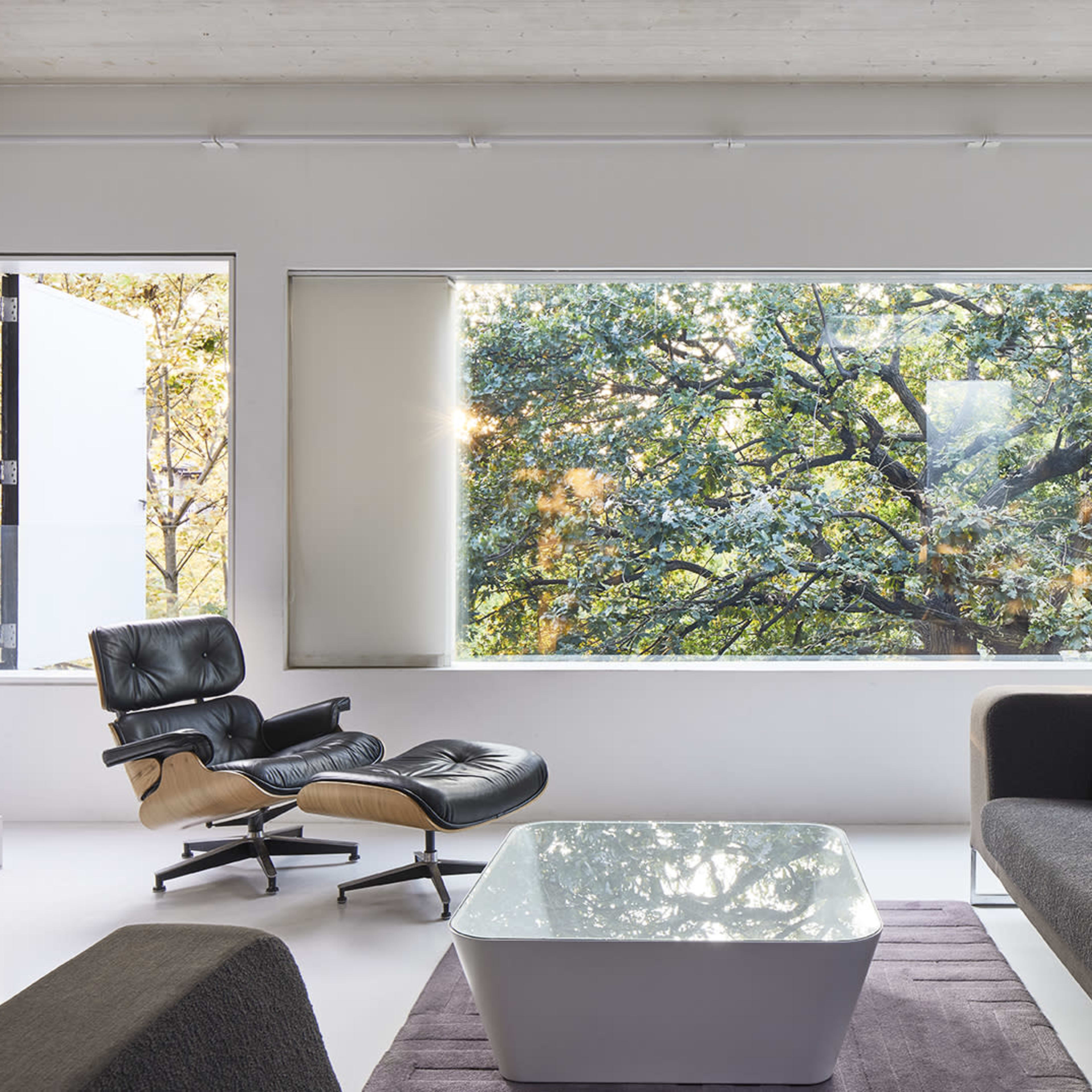 A modern living room featuring a black and beige lounge chair, a white coffee table, and a large window displaying a view of green trees outside.