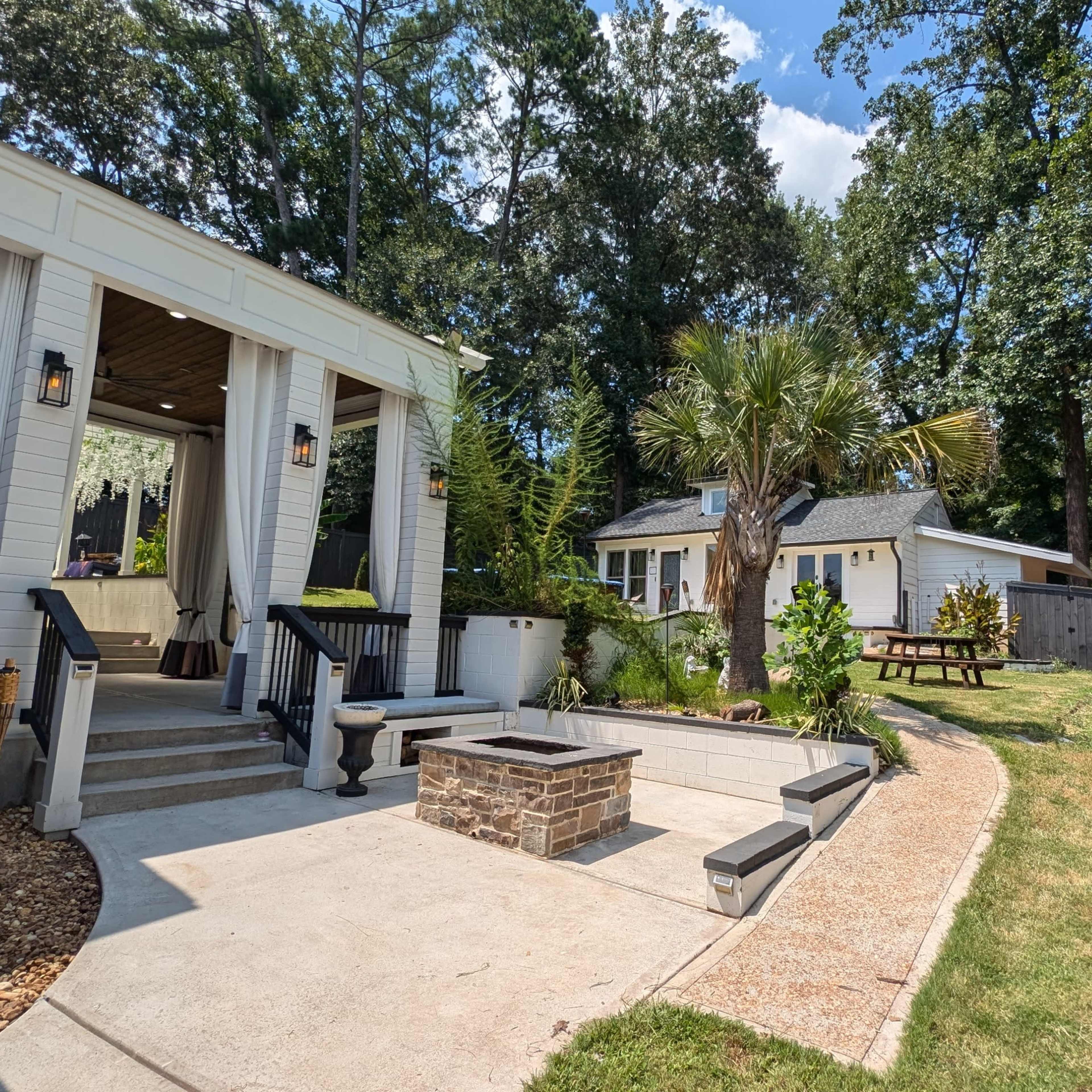 A backyard area with a patio featuring steps and a fire pit, surrounded by a lawn and play equipment, alongside a house and another structure in the background.