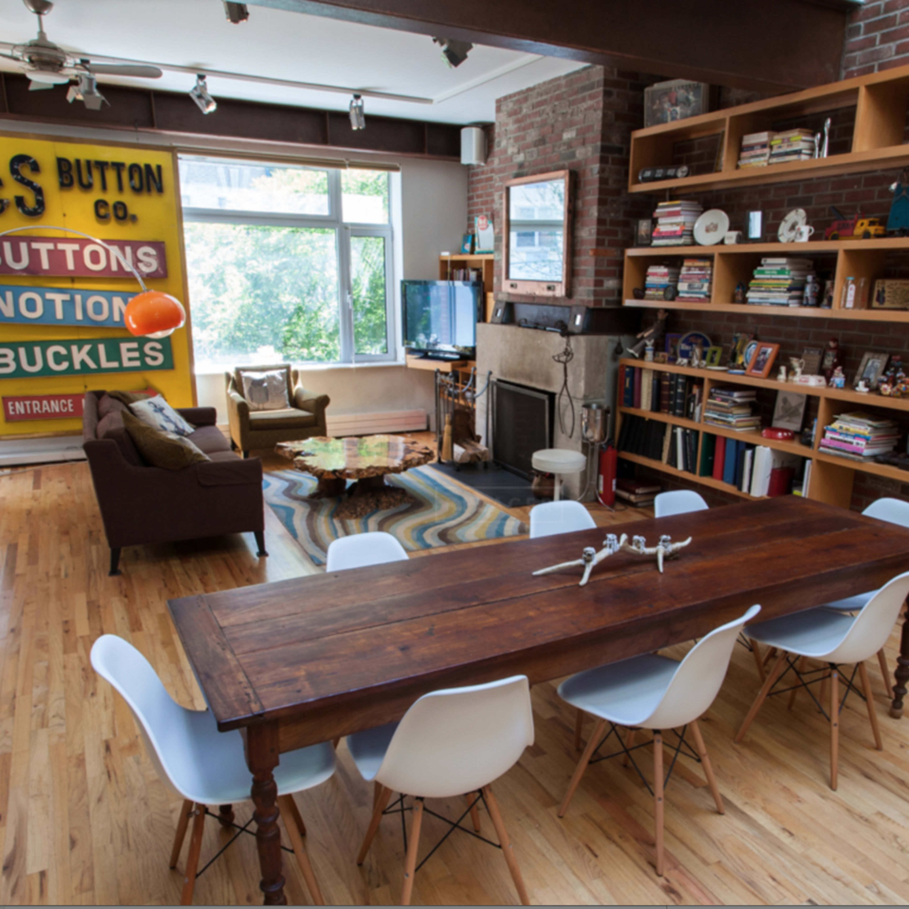 A spacious living area featuring a wooden dining table surrounded by white chairs, with a brick wall and shelves filled with books and decorative items.