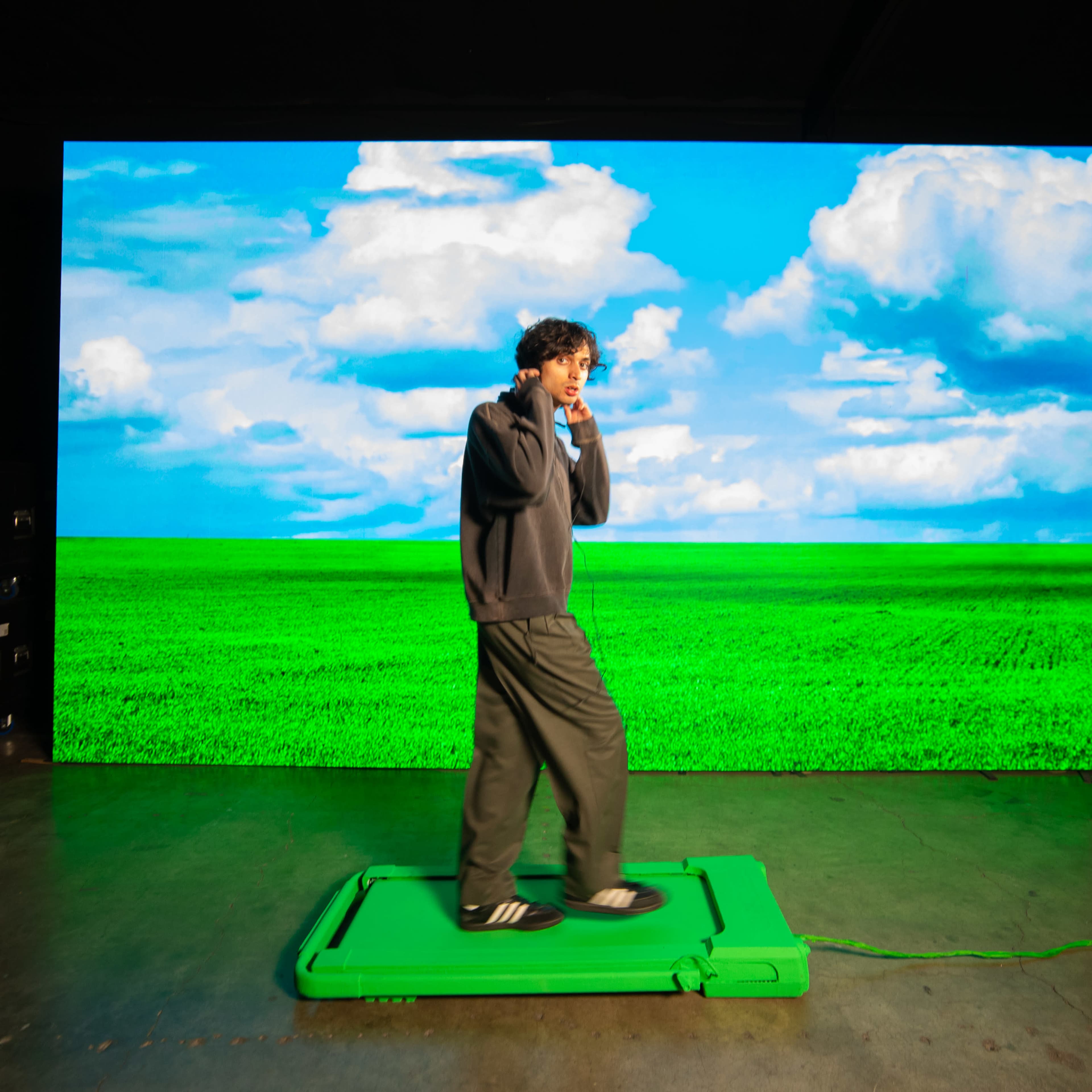 A person stands on a green platform in front of a large screen displaying a blue sky and green field.