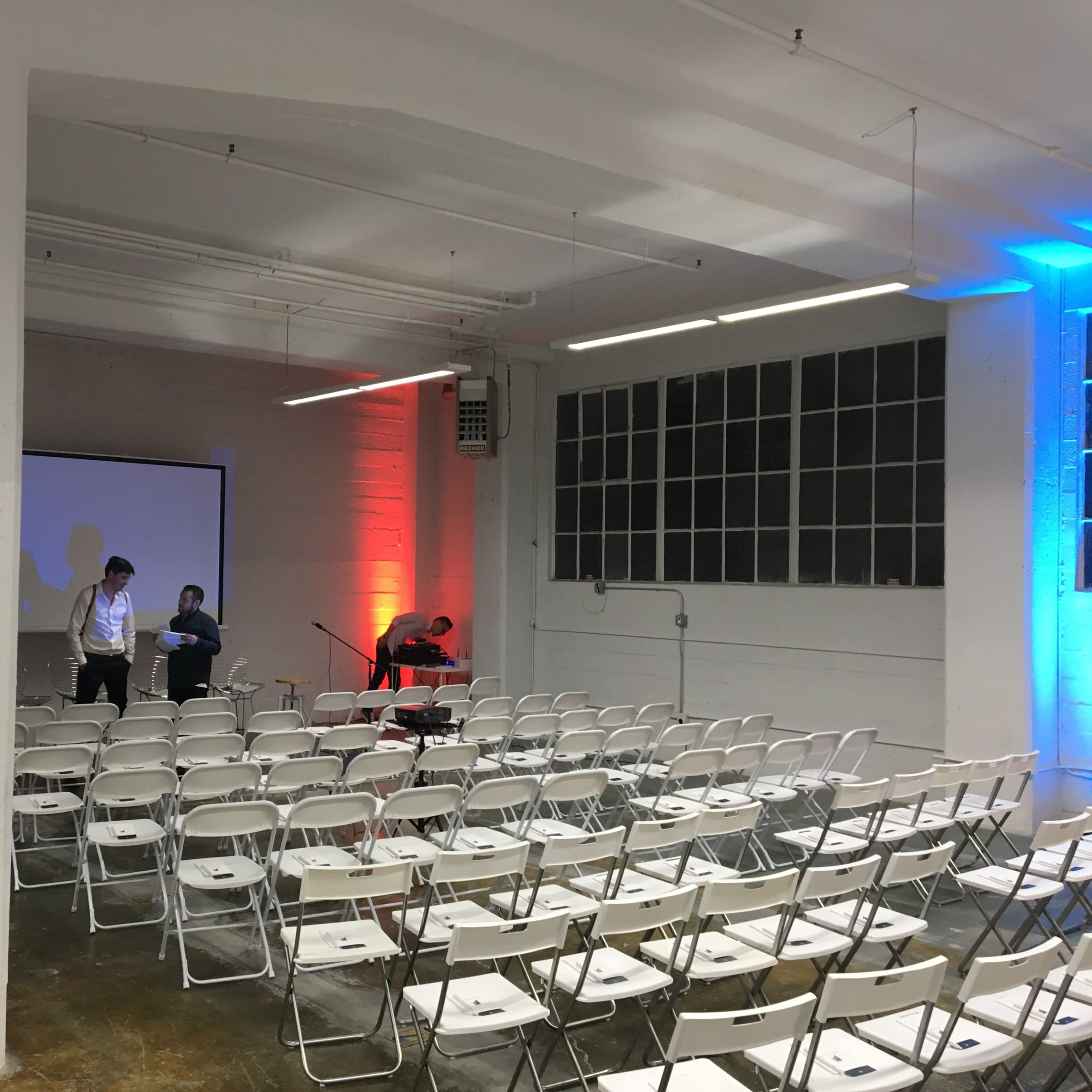 A spacious, empty room with rows of white chairs arranged for an event, featuring a projector screen and ambient lighting in red and blue.