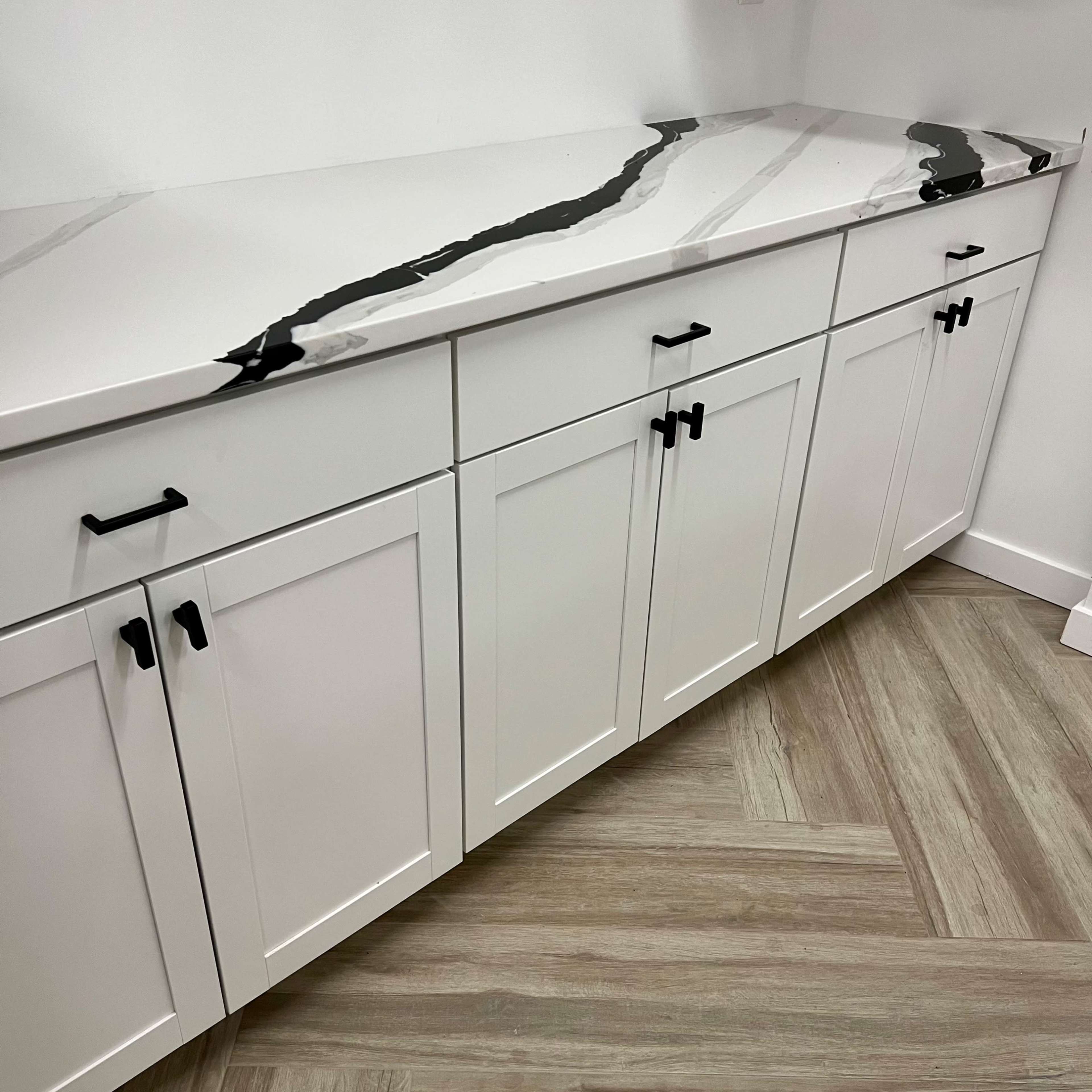 A white cabinet with black handles and a patterned marble countertop, set against a light-colored wall and floor with a wood-like texture.