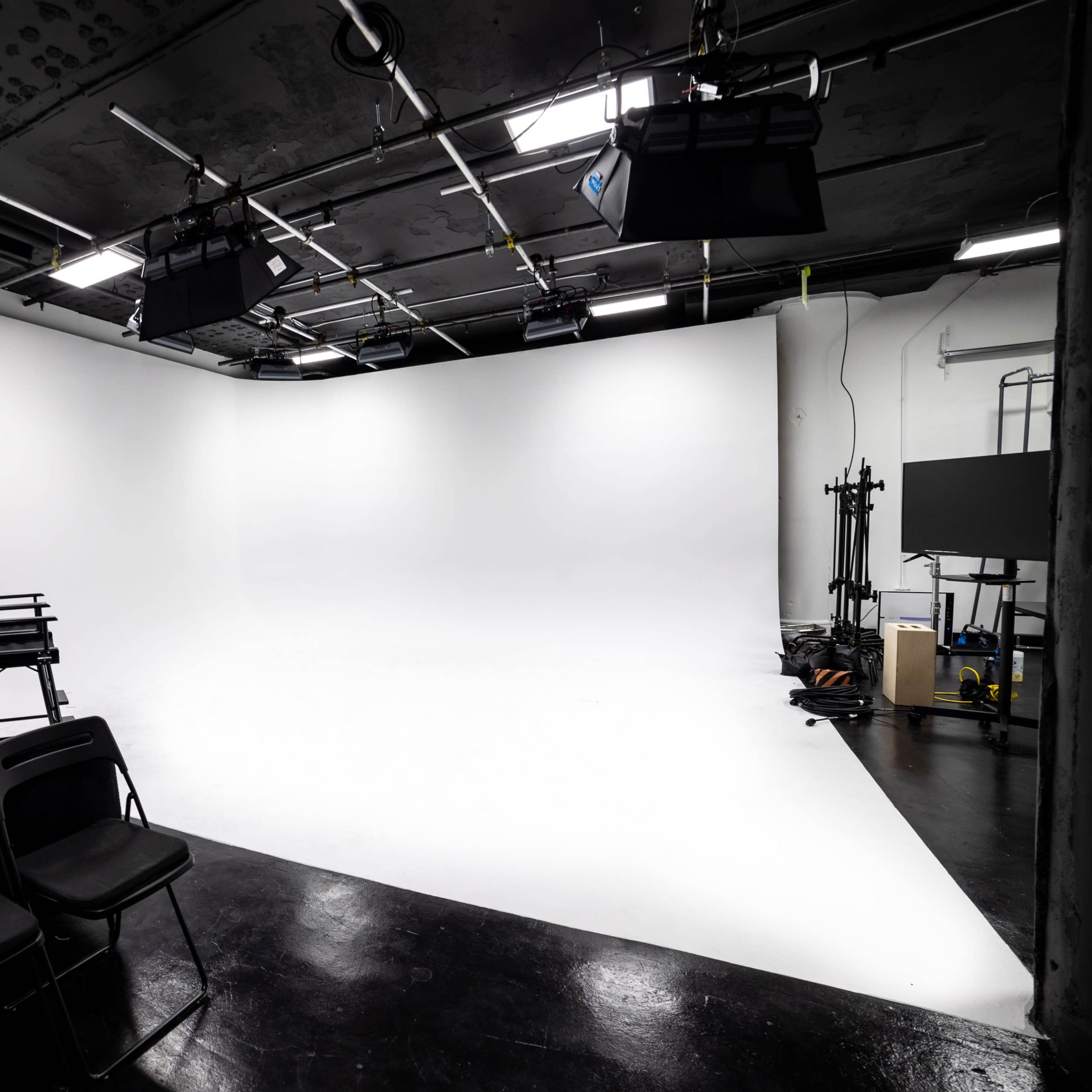 A photography studio featuring a large white backdrop, studio lighting, a mirror with lights, a director's chair, and several black folding chairs.