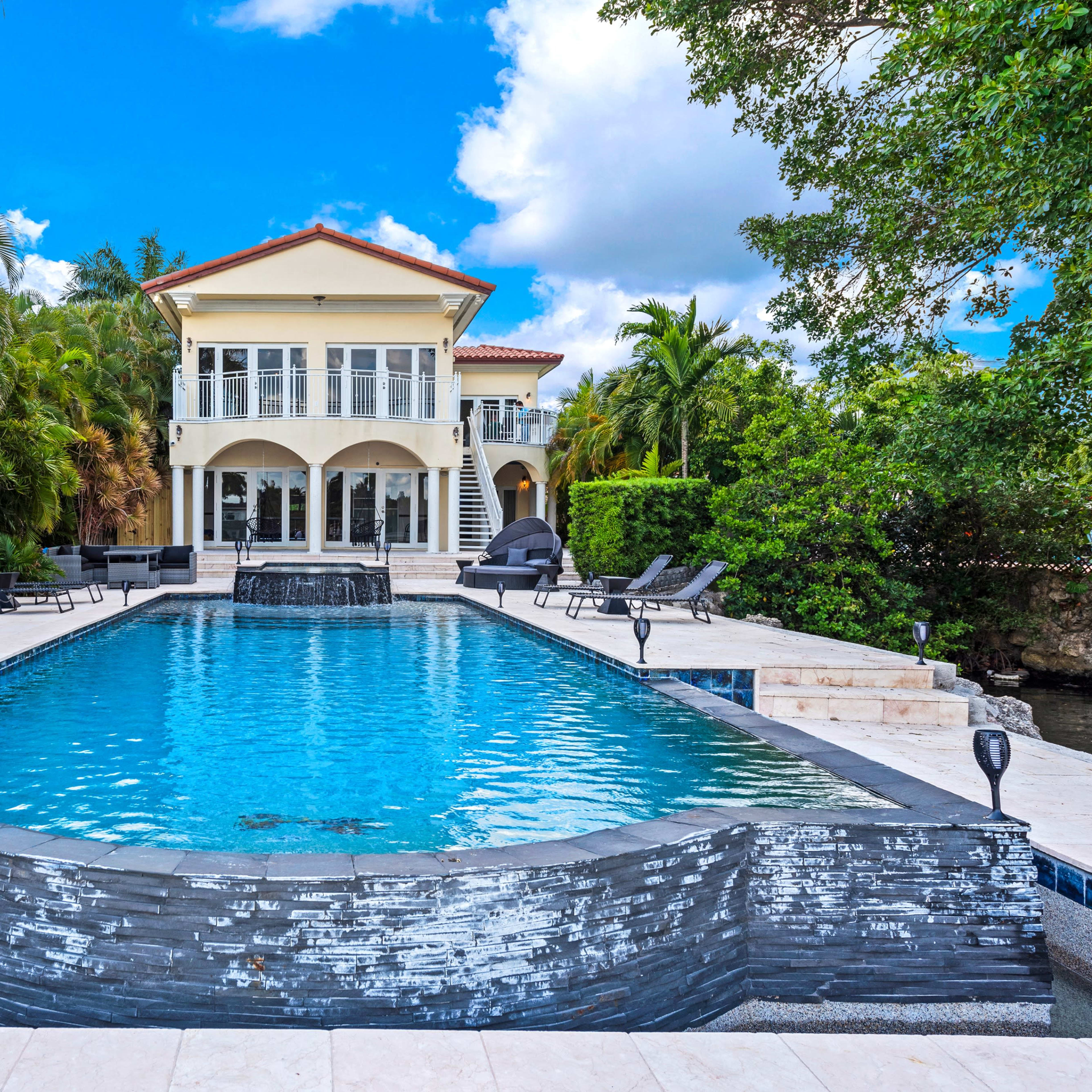 A spacious swimming pool with a waterfall feature in front of a multi-story house surrounded by tropical landscaping.