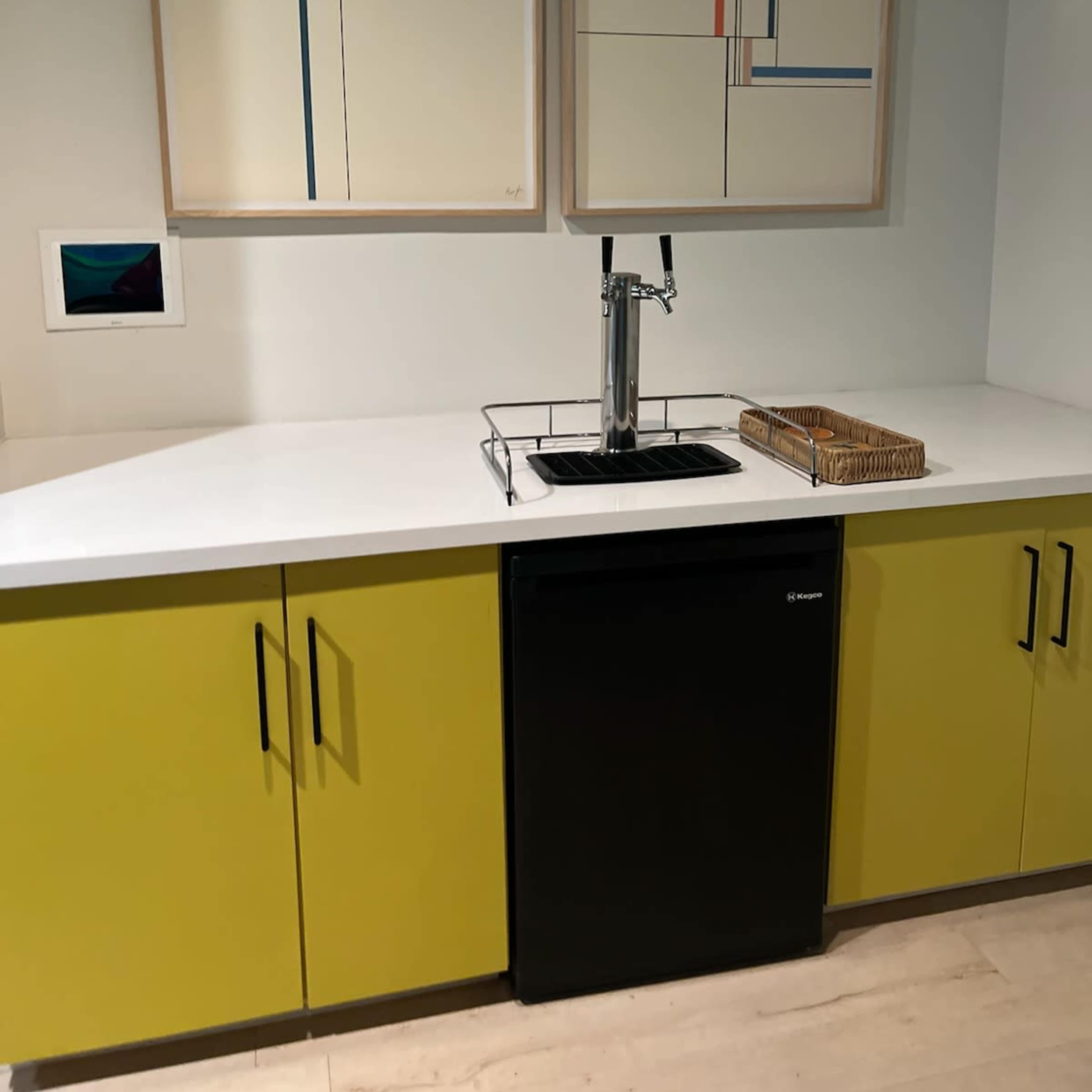 A modern kitchen area featuring a yellow cabinet with a black fridge, a white countertop, and two abstract artworks hanging on the wall above.