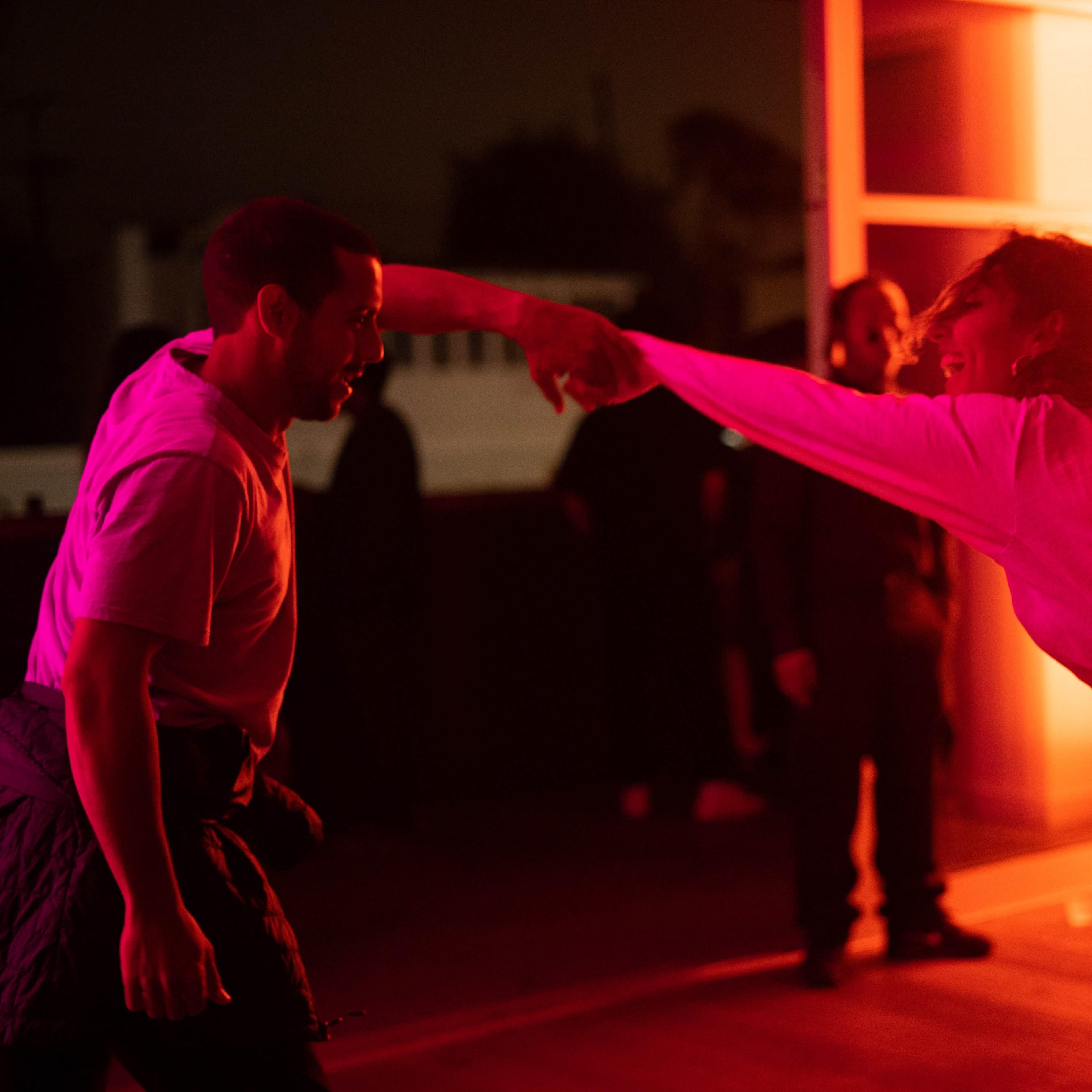 Two people are dancing in a dimly lit room with vibrant red lighting, while several onlookers watch in the background.