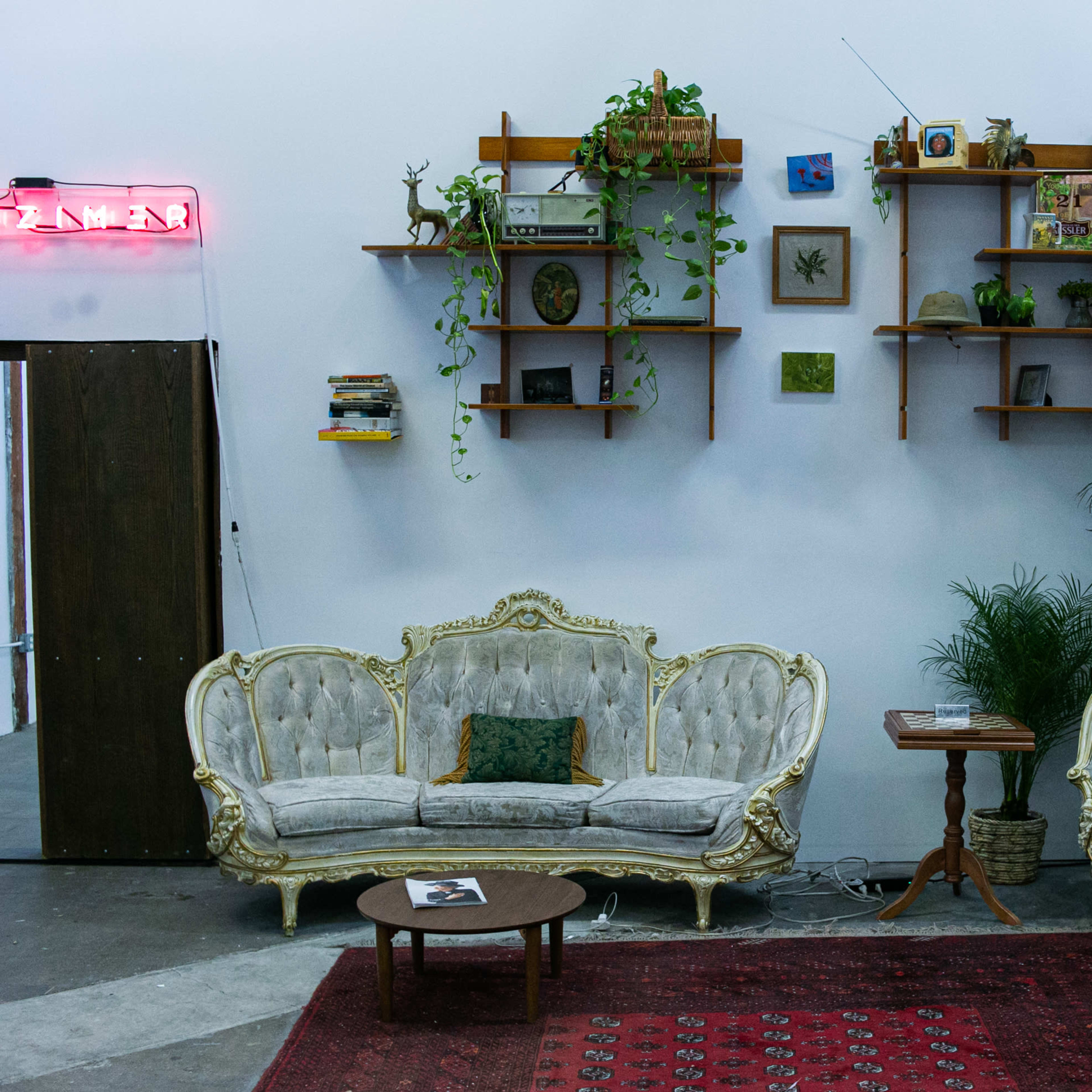 A vintage-style seating area with two ornate sofas, a small table, and shelves adorned with plants and decor items against a white wall.