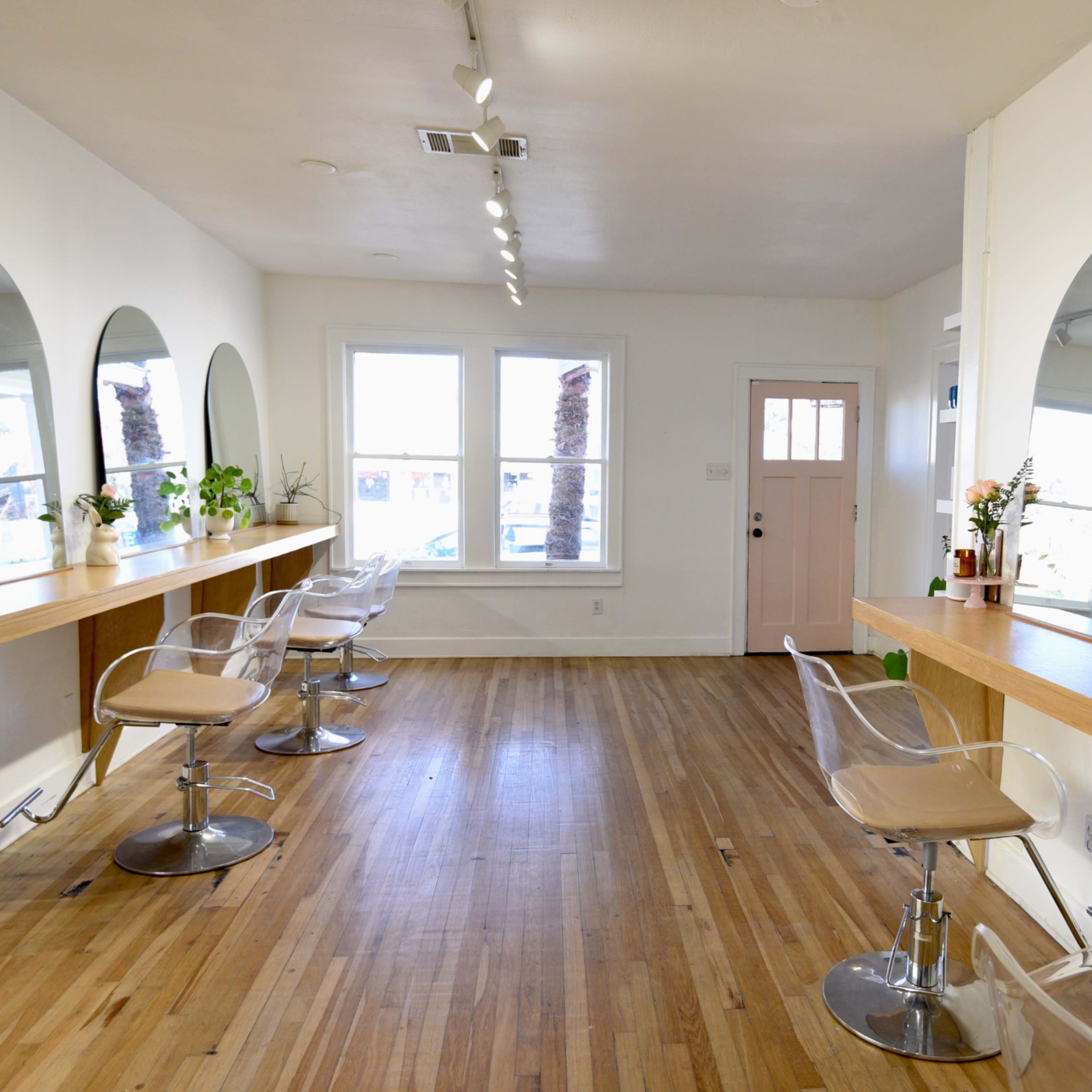 A well-lit salon with three mirrored styling stations and clear chairs, featuring wooden counters and large windows.