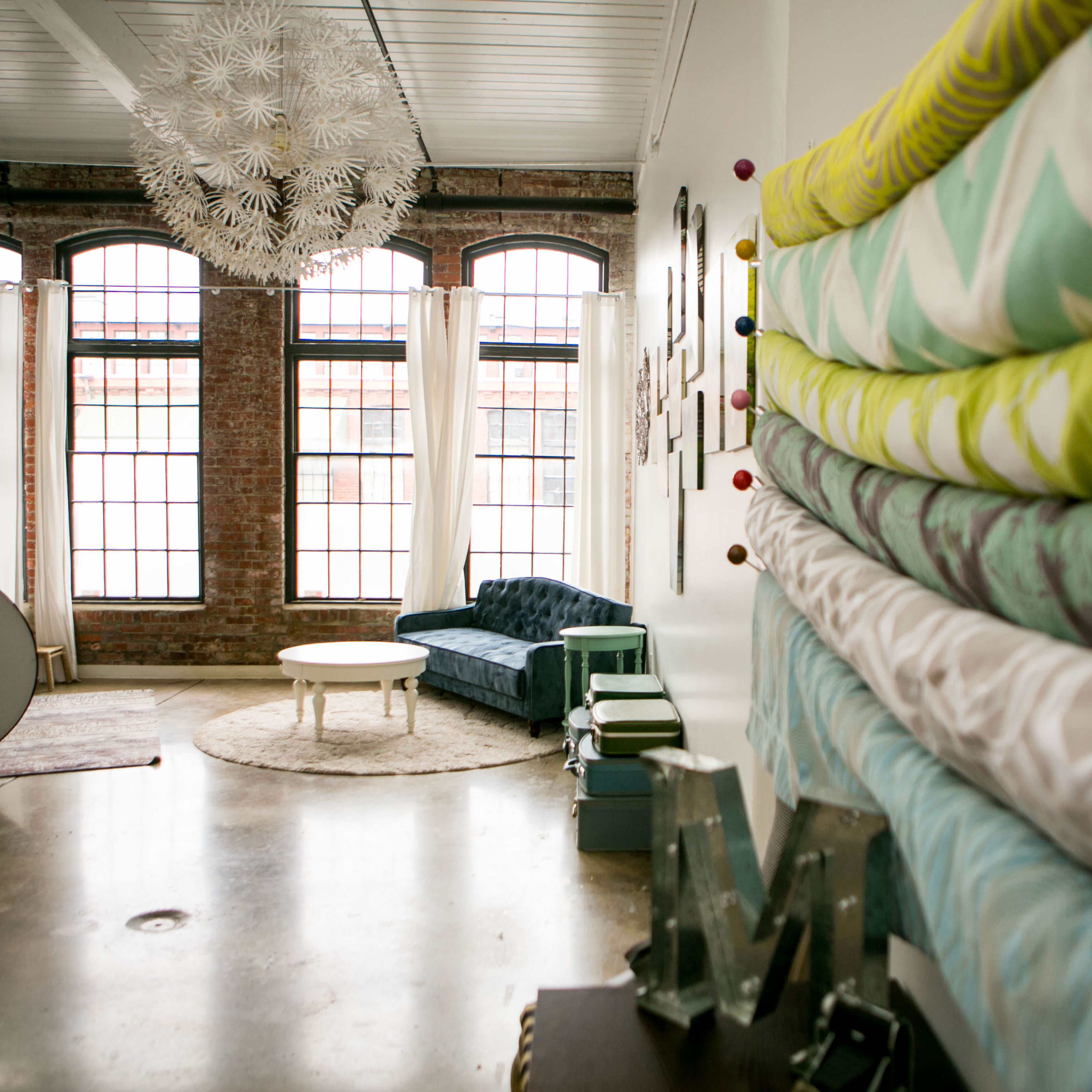 A bright, spacious interior with large windows, a blue couch, and a stack of colorful fabric rolls against the wall.