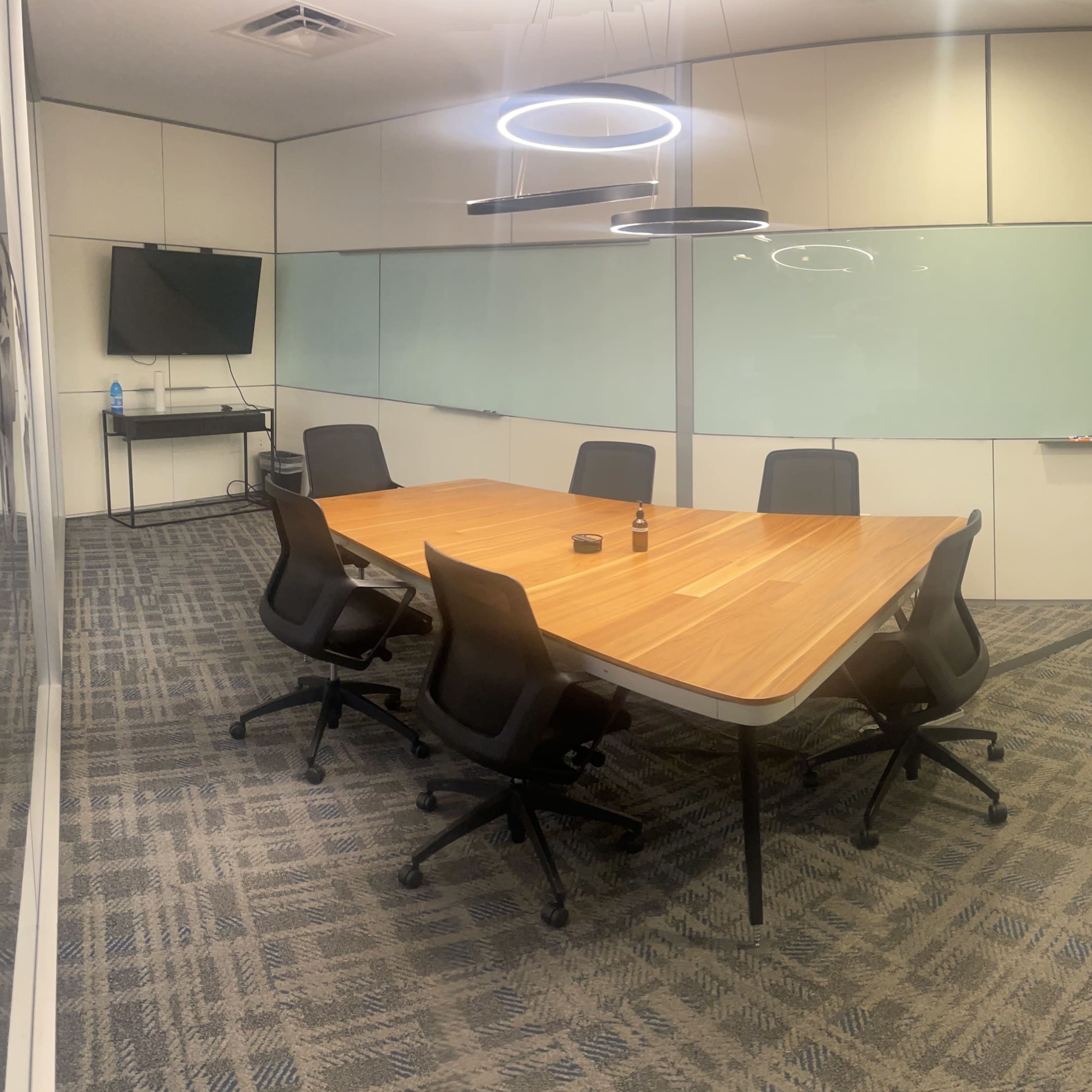 A modern conference room features a large wooden table surrounded by six ergonomic chairs, with a television and glass walls.