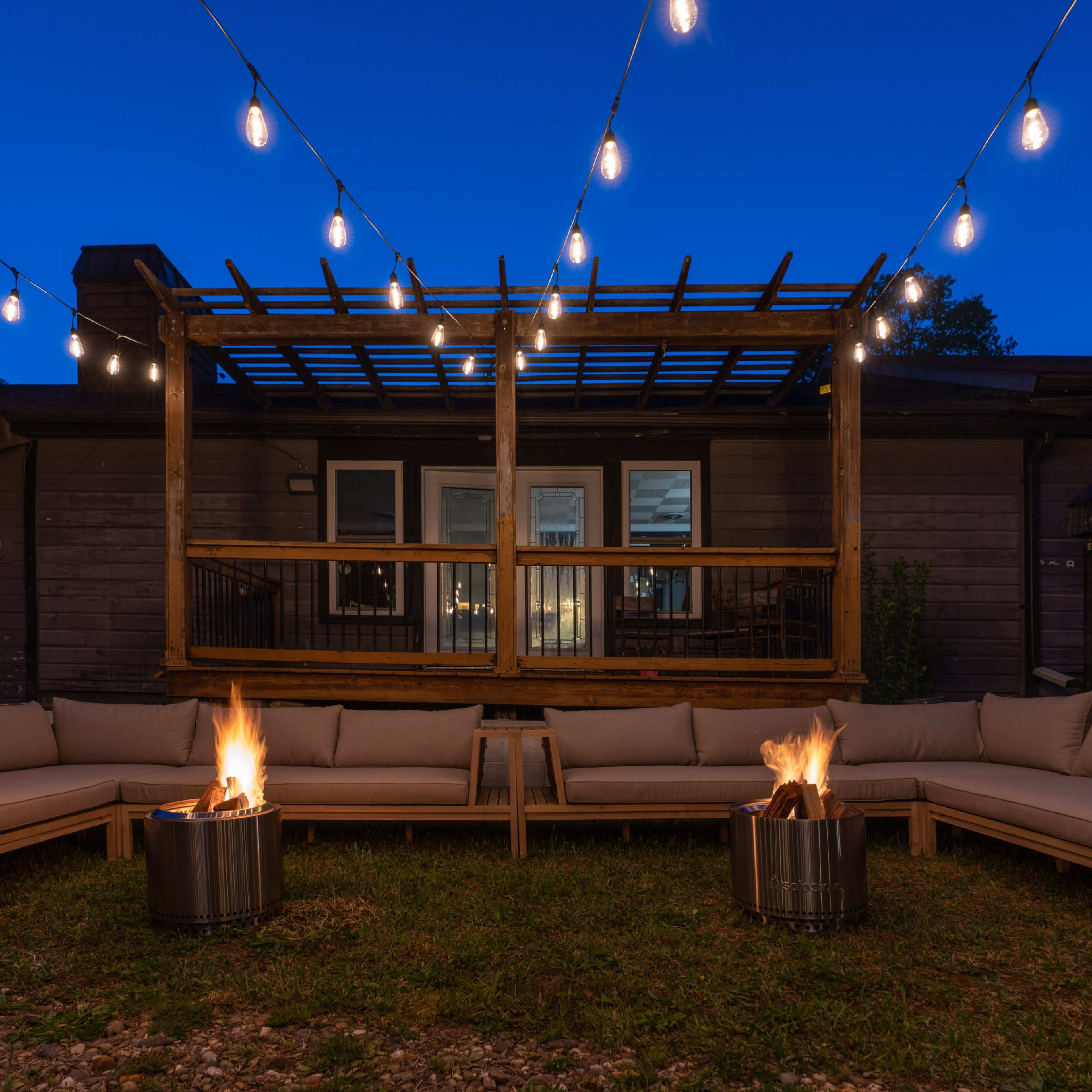 An outdoor seating area with two fire pits on a lawn, surrounded by a large sectional sofa and strung lights overhead at dusk.
