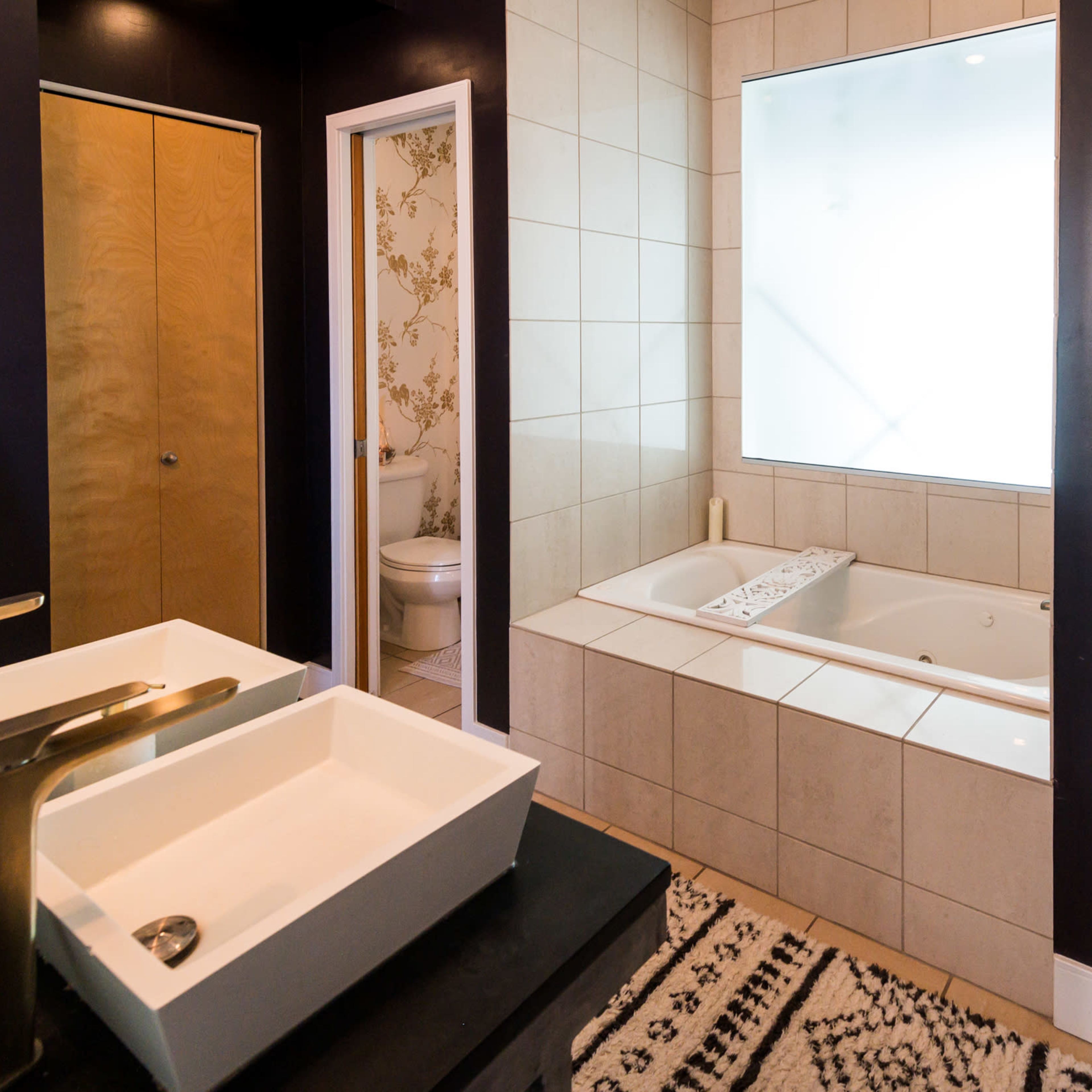 A modern bathroom featuring a large window above a jetted bathtub and dual sinks with a dark accent wall.