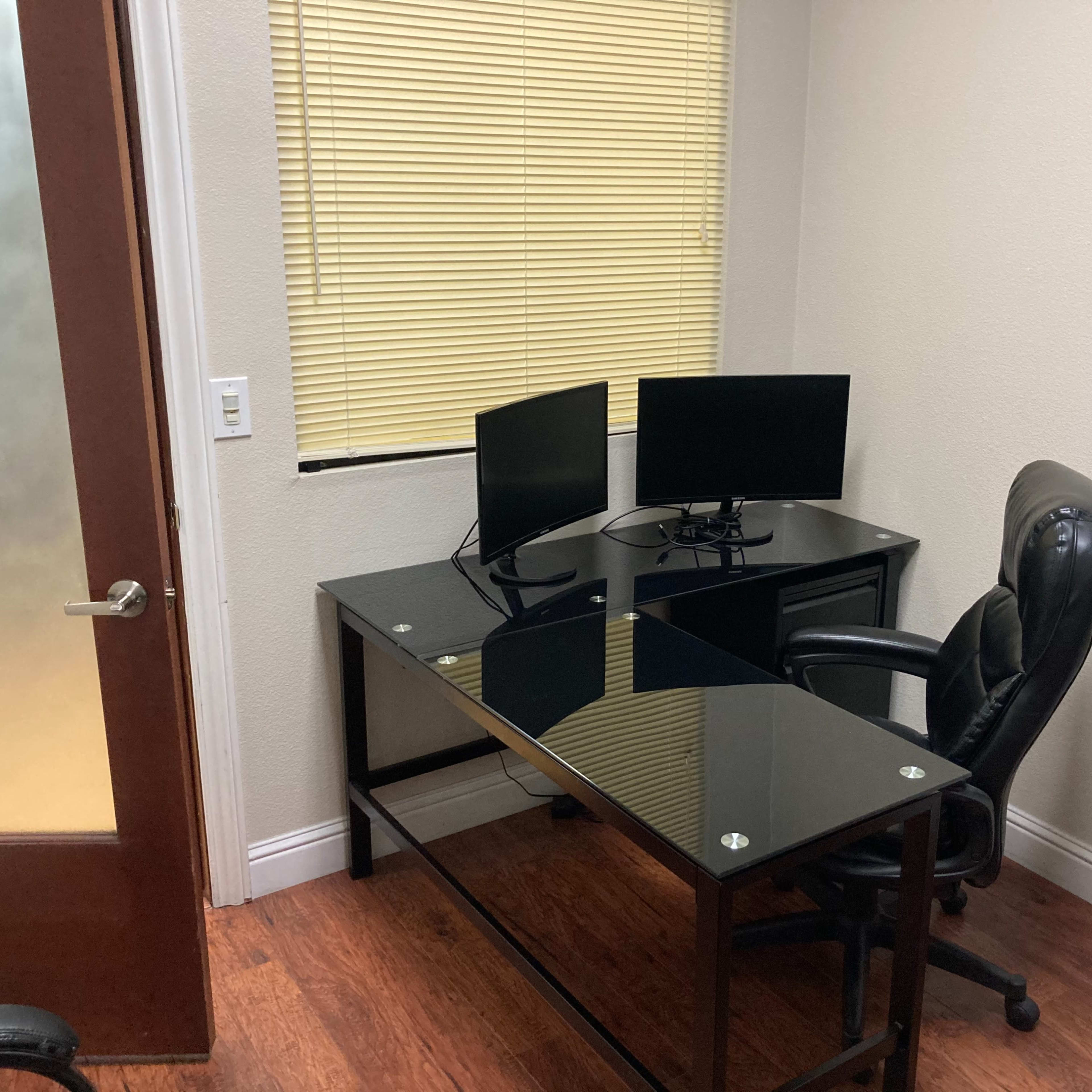 A small office with a glass-top desk, two computer monitors, a black leather office chair, and a window covered with blinds.
