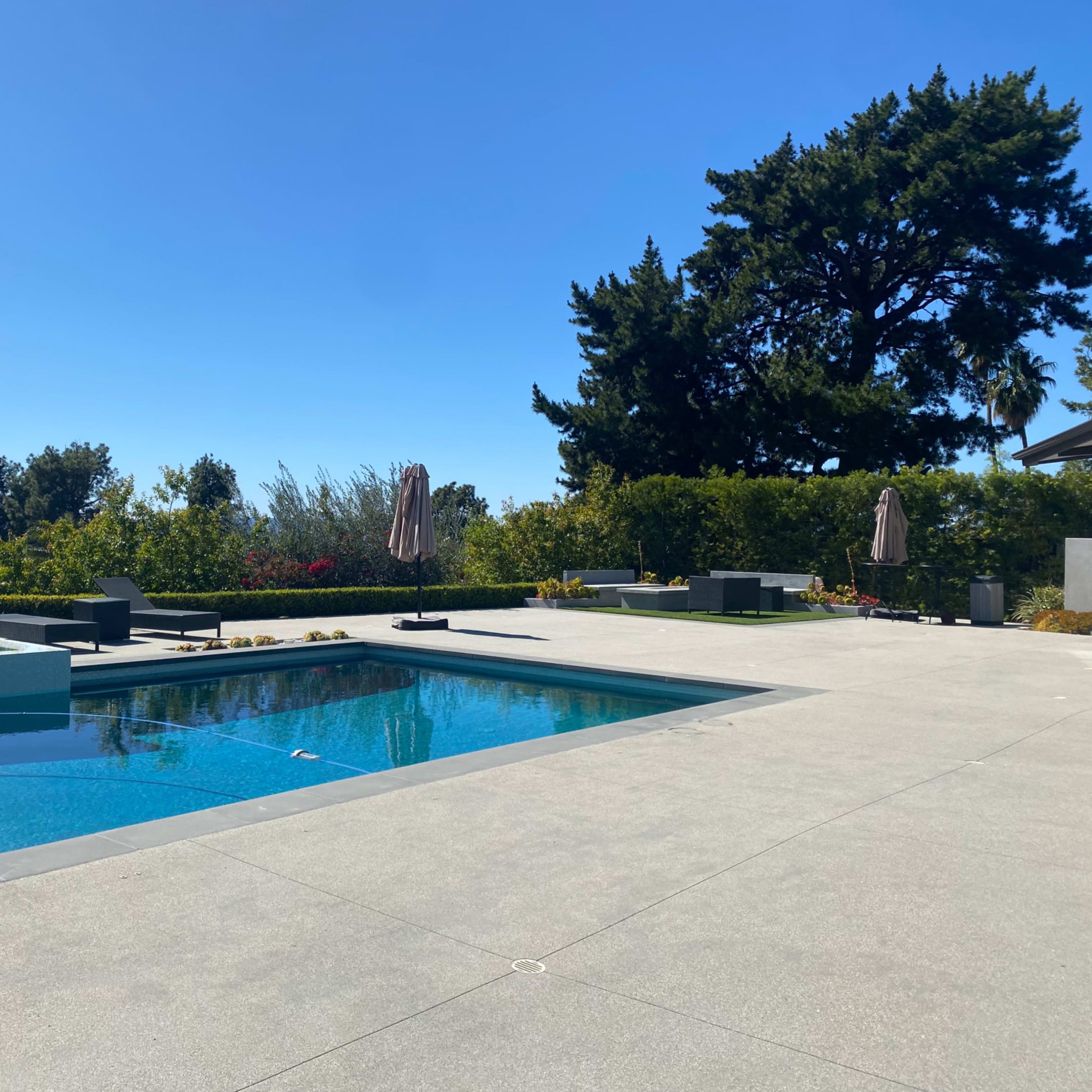 A modern outdoor pool area features a clear blue pool, lounge chairs, and landscaping under a bright blue sky.