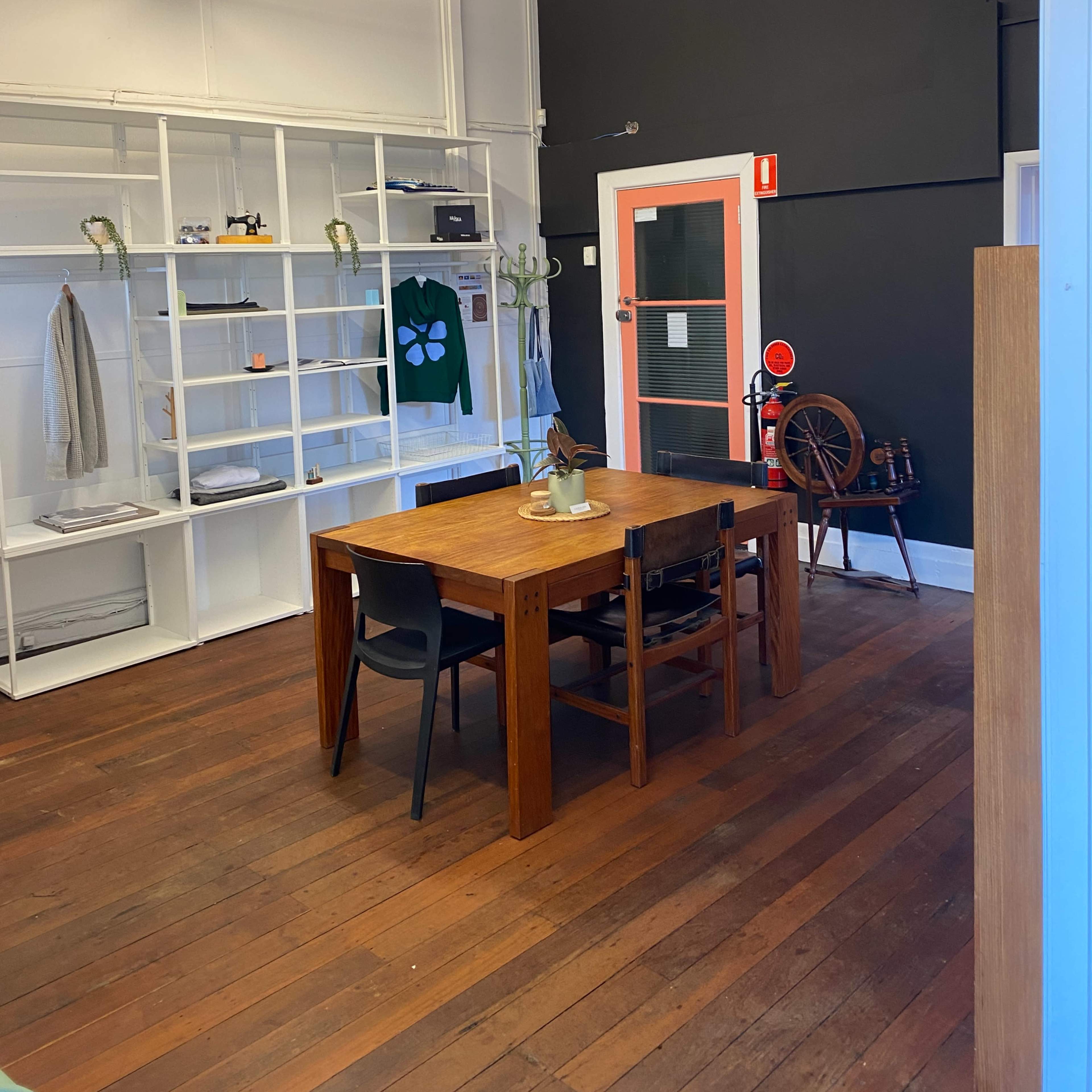 An office space with a wooden table surrounded by black chairs, shelves on the walls displaying various items, and a black wall contrasting with a bright orange door.